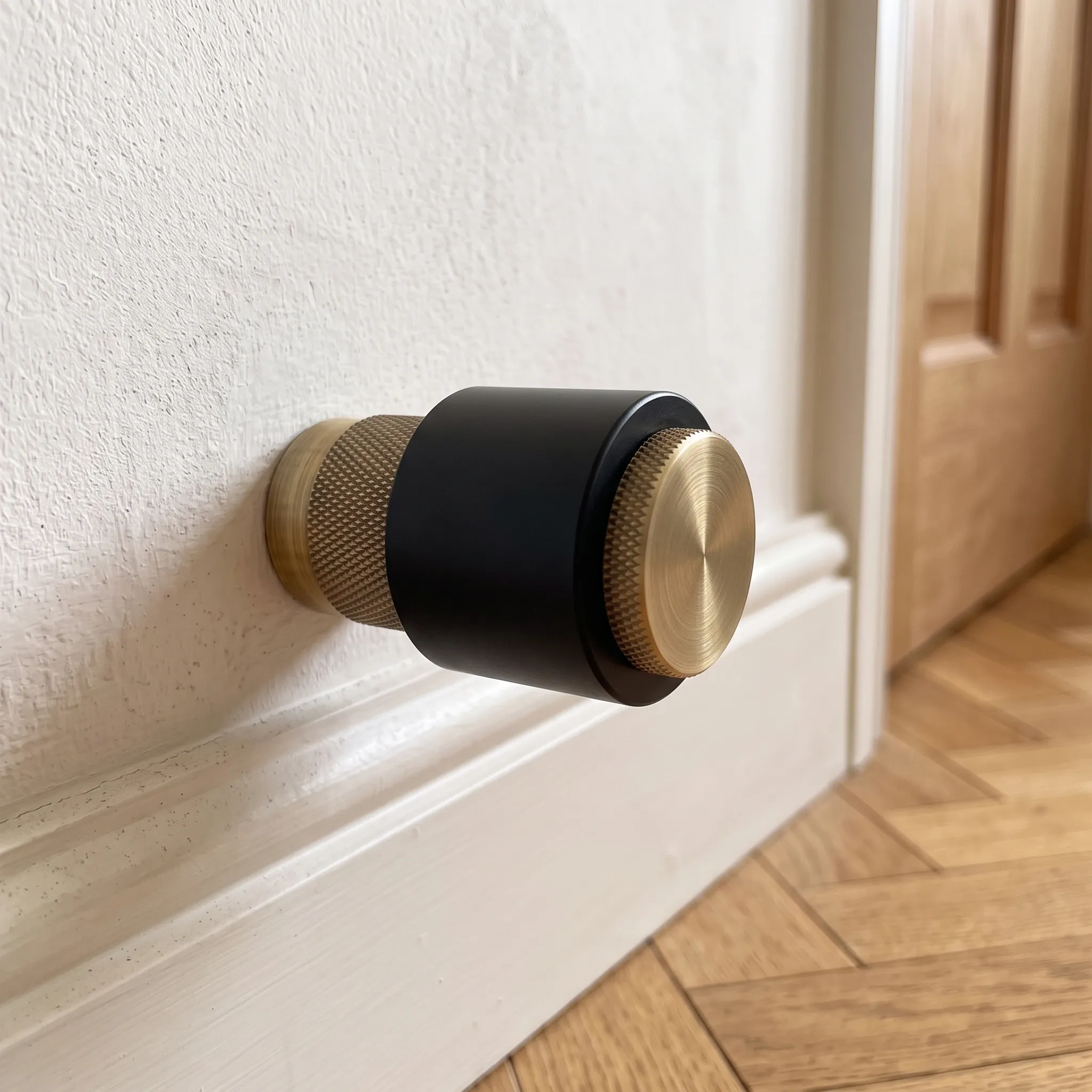 The Brass Gold knurled door stop photographed in a bright interior beside an open wooden door with herringbone parquet flooring.