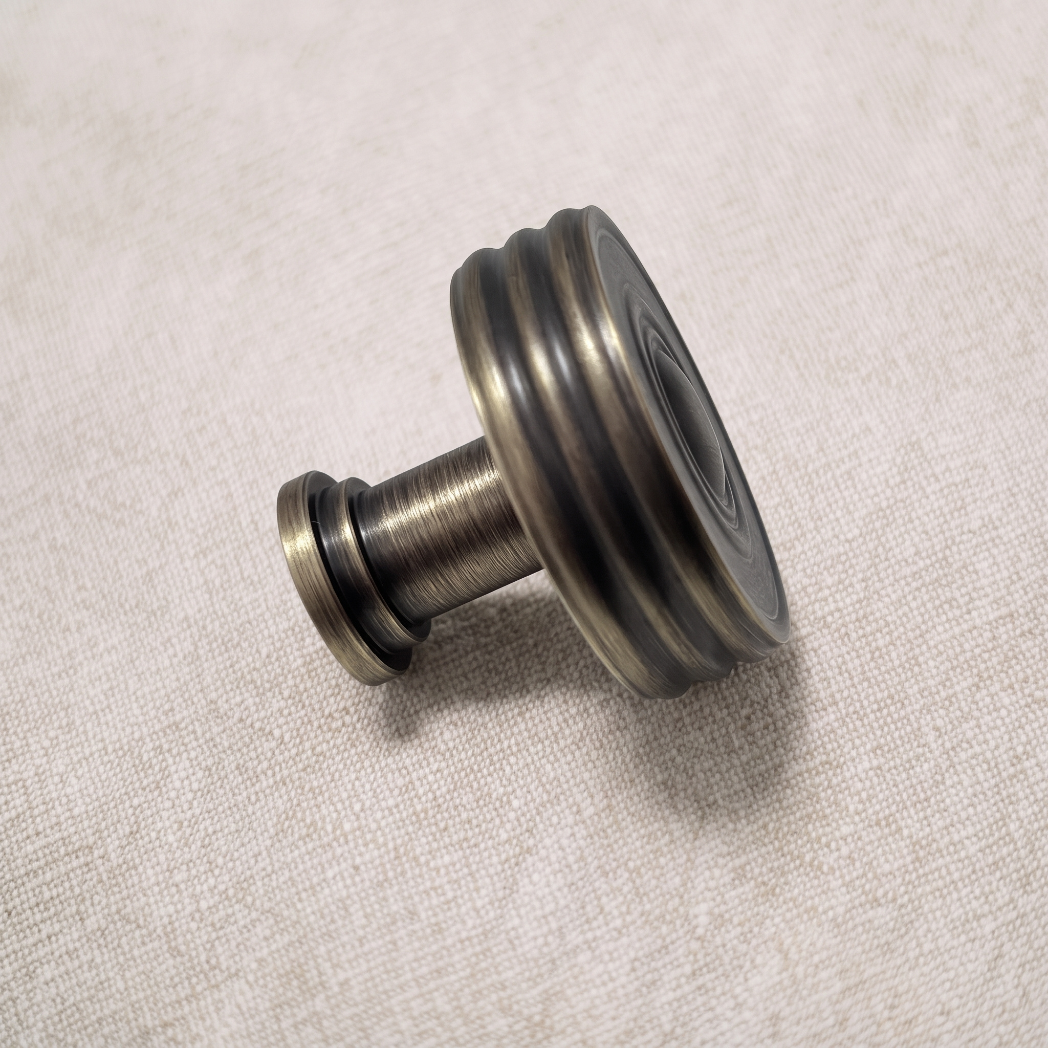 Side view of the antique bronze circular knob resting on pale linen, showing its stacked bands, neck and base collar