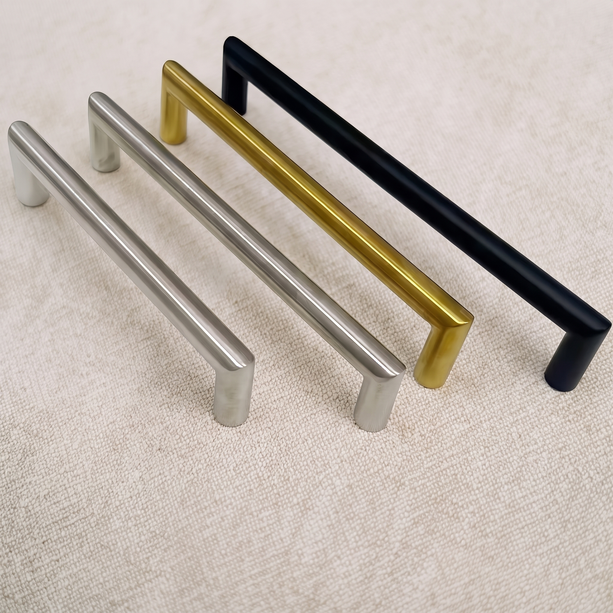 Four T-bar cabinet pulls from the range arranged on pale linen, spanning brushed nickel, brush gold and matt black finishes with softened rectangular bars