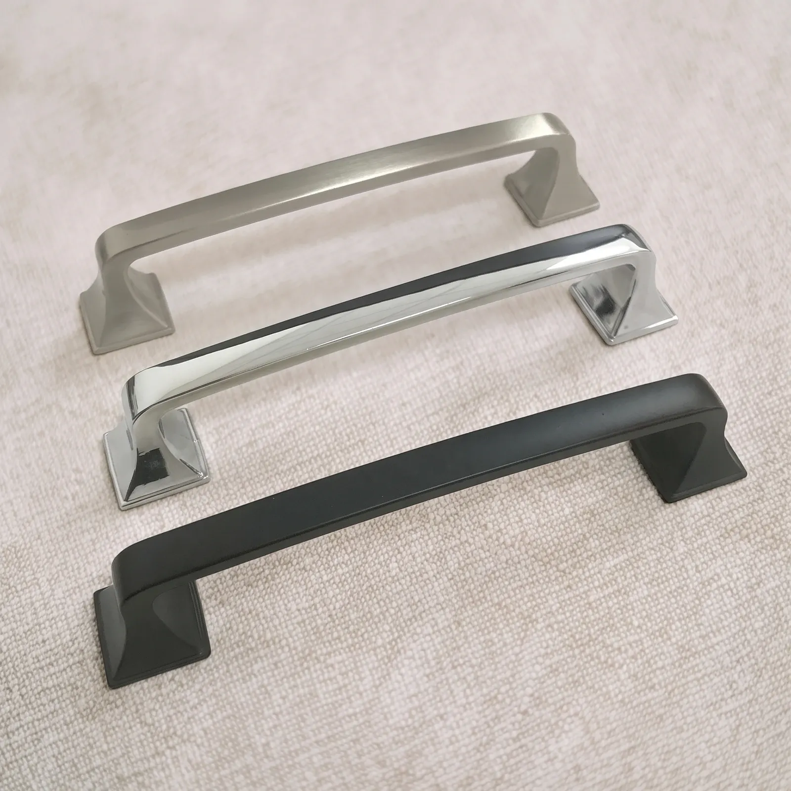Angled view of three ZAMAK arched cabinet pulls arranged diagonally in Brushed Nickel, Chrome and Black, each with a crowned bar, faceted pyramid feet and square base plates