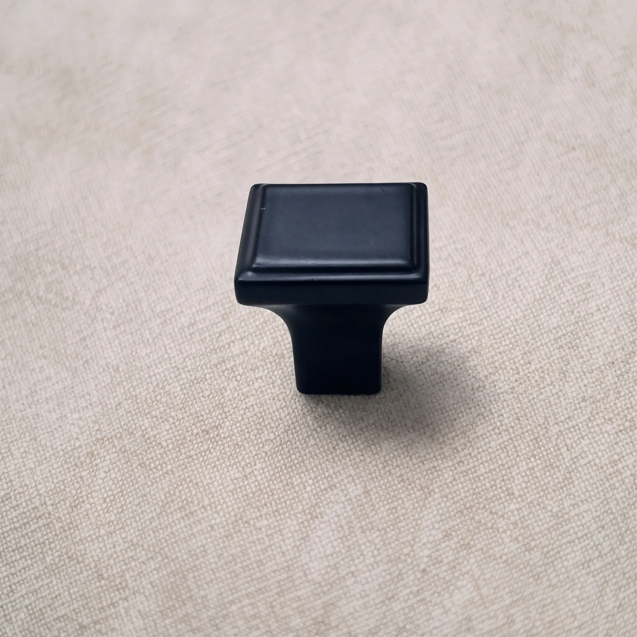 Top angled perspective of a Matt Black square cabinet knob displaying its stepped rim and flared pedestal on a textured beige studio surface.