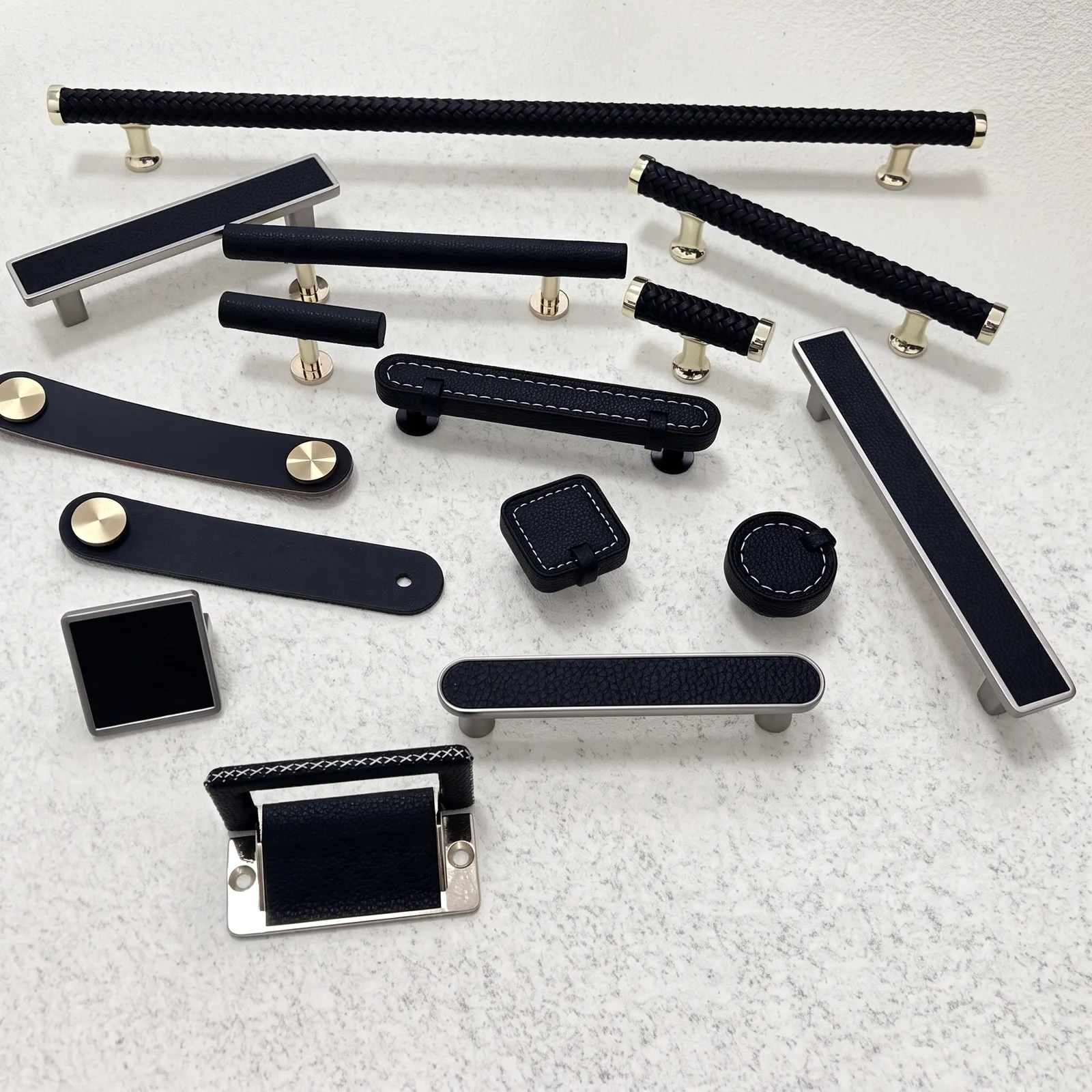 Angled view of black leather strap pulls with gold-toned rivets beside framed and stitched leather knobs on a pale textured surface.