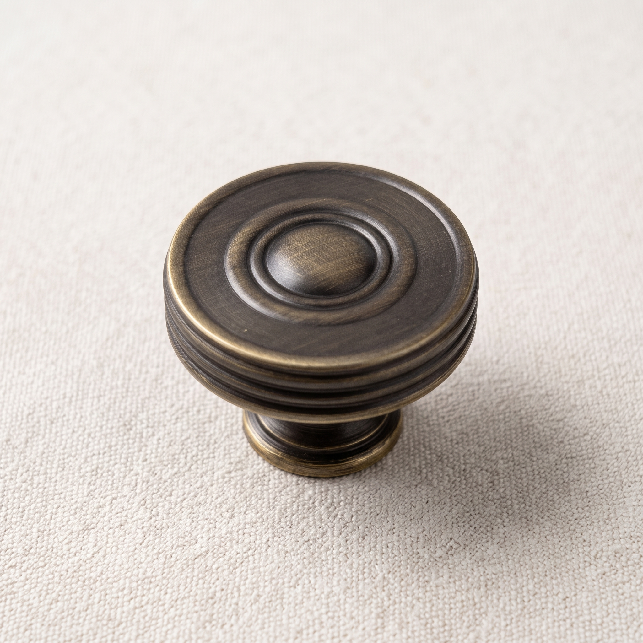 Angled overhead view of the antique bronze 32 mm circular knob on pale linen, showing concentric bands around a raised centre and the banded side