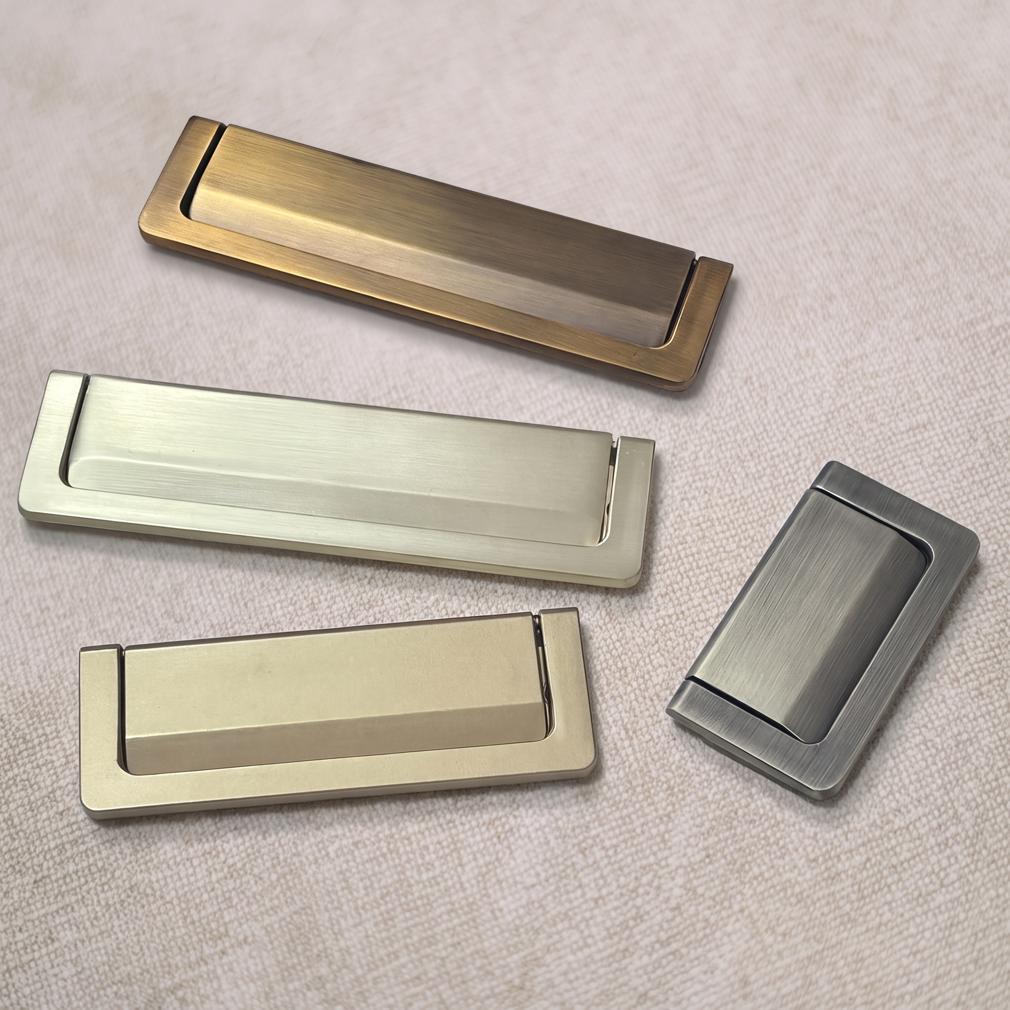 Four recessed flip pulls in aged brass, pale champagne gold and brushed nickel, set at angles on a pale beige textured surface.