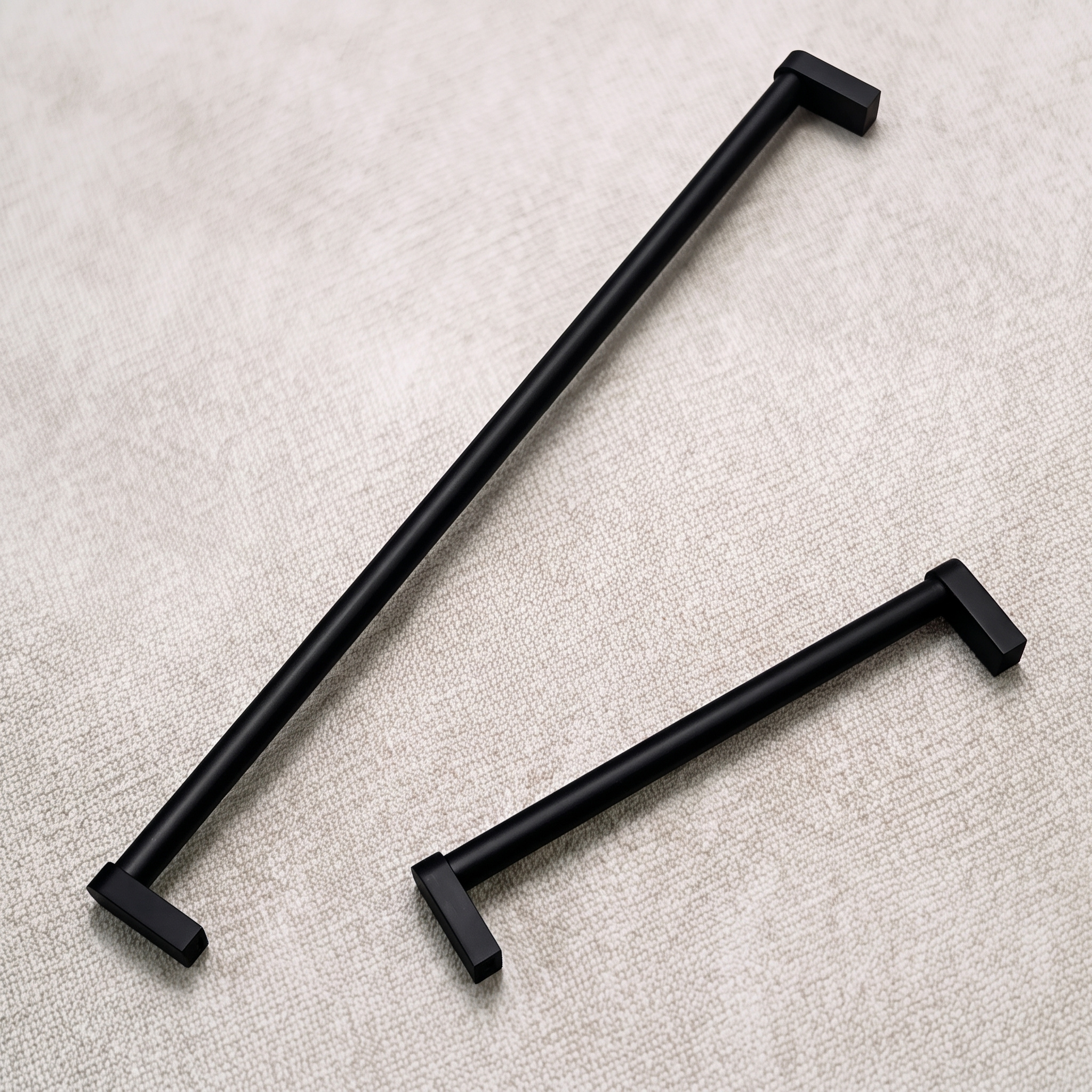 Two matt black smooth cylindrical aluminum bar pulls of different lengths arranged side by side on pale linen