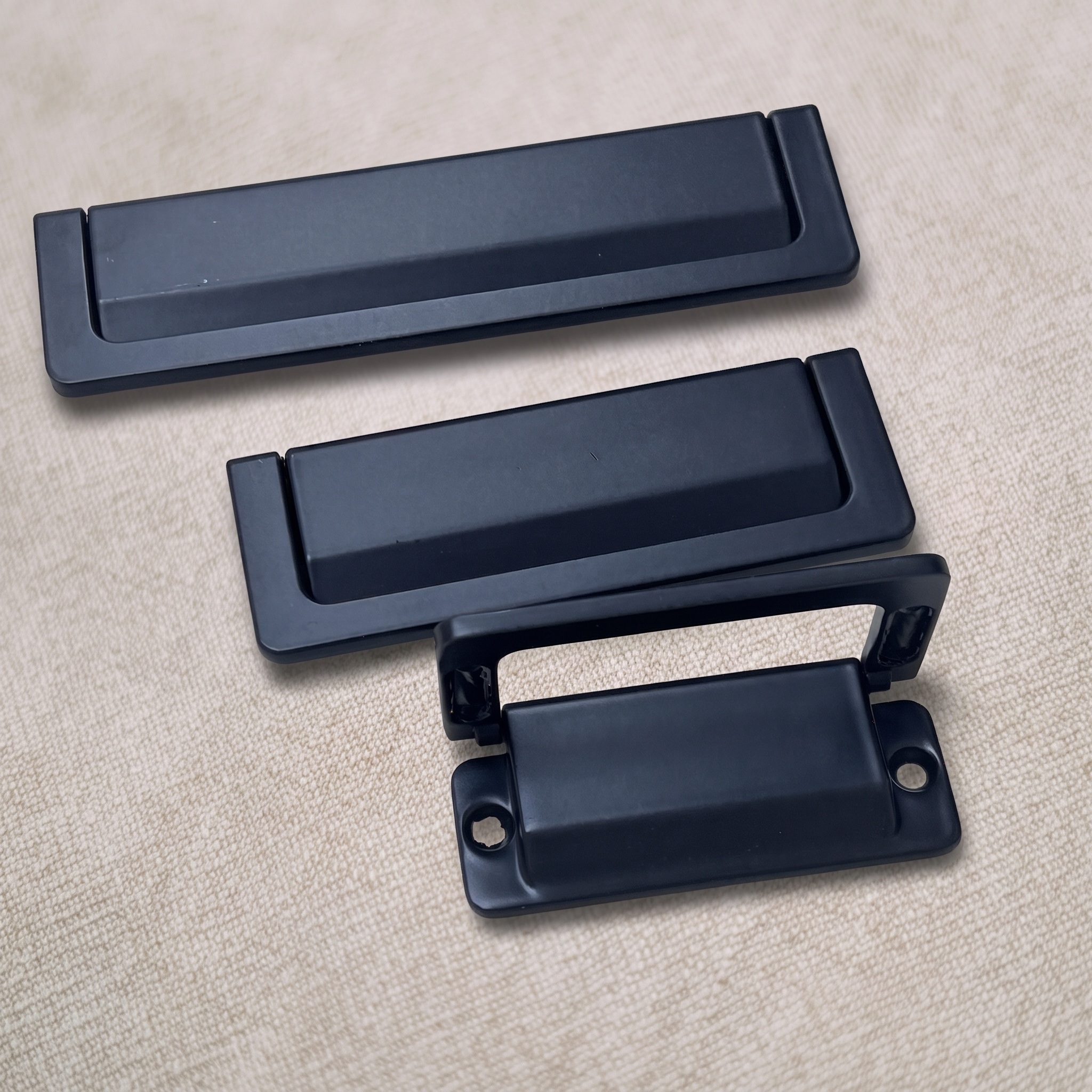 Three matte black recessed flip pulls on a pale beige textured surface, shown closed, part-lifted, and fully open with the rear mounting plate and two screw holes.