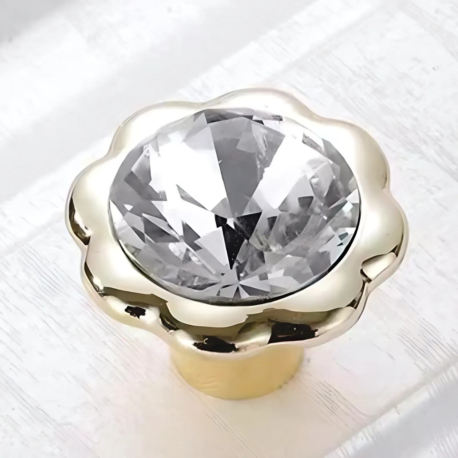 Angled view of one round faceted clear crystal knob framed by a scalloped gold-tone rim on a gold-tone base, standing on white painted wood.