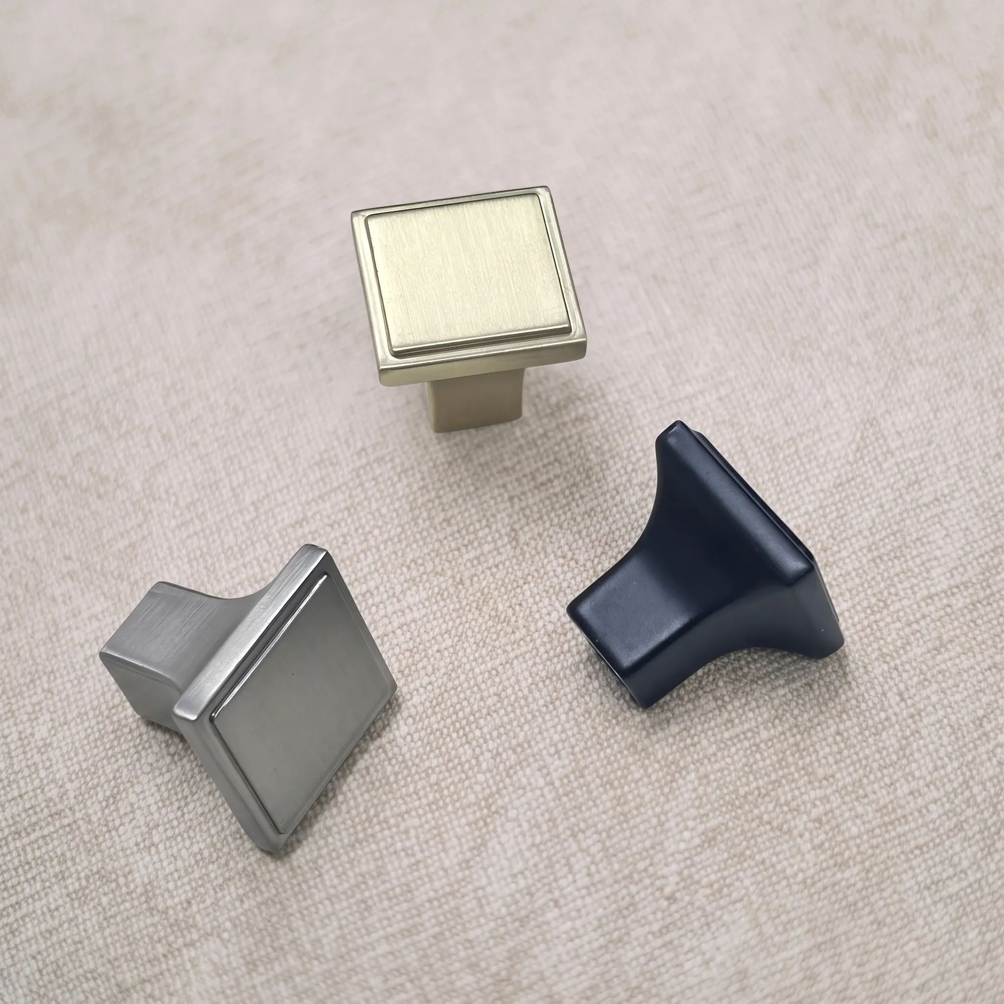 Elevated view of Brush Gold, Satin Nickel, and Matt Black square knobs, with tilted views showing flared pedestals on a textured beige surface.