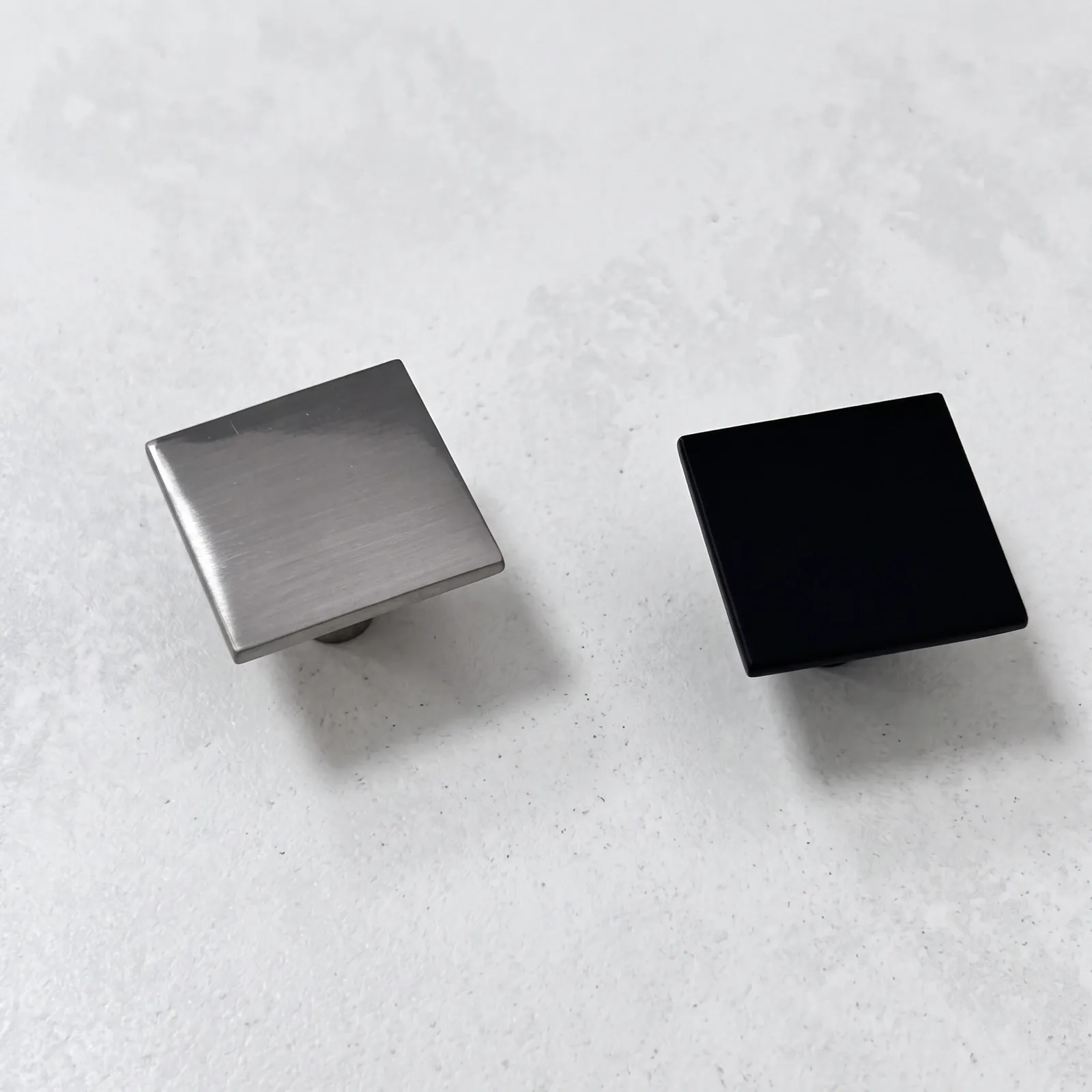 Brushed nickel and matt black square flat-plate cabinet knobs side by side on a pale ground.