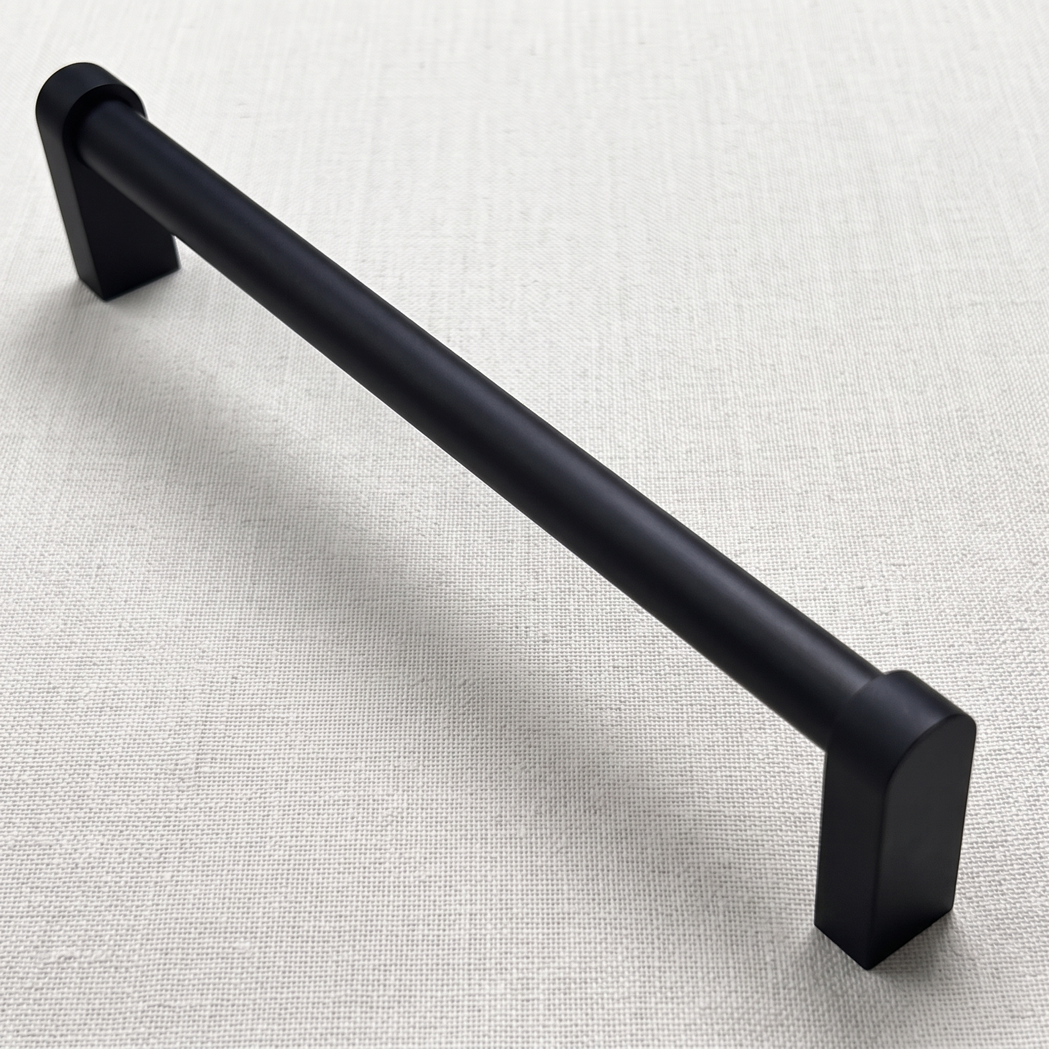 Angled view of a matt black smooth cylindrical aluminum bar pull on pale linen, showing domed collars joining the round bar to square block feet