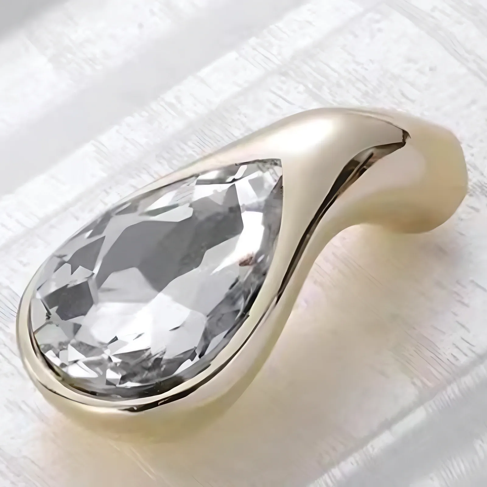Angled view of a teardrop drop pull featuring a pear-shaped faceted clear crystal in a curved gold-tone mount, on white painted wood.