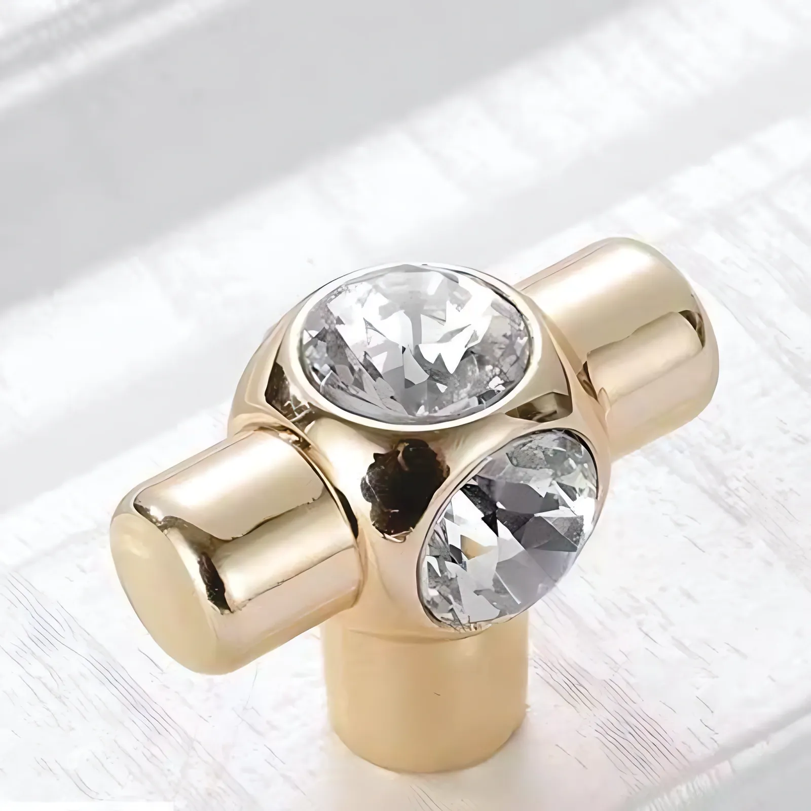 Angled view of a T-shaped gold-tone knob carrying two round faceted clear crystals, standing on white painted wood.