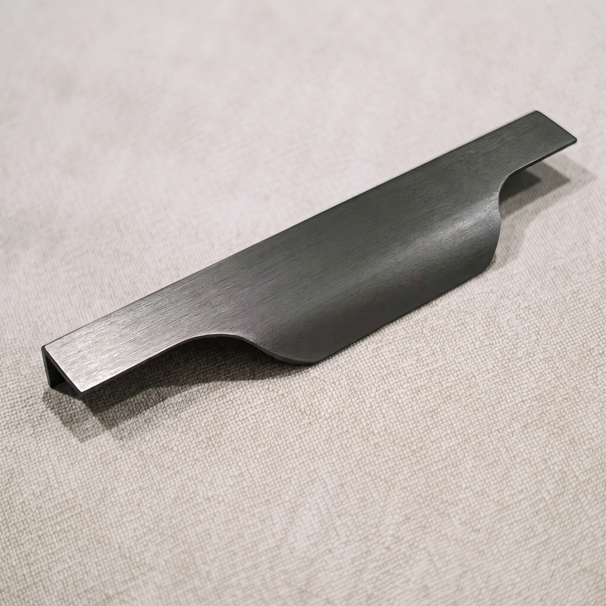 Angled view of a dark gunmetal-toned aluminum overlay edge pull on a pale linen surface, showing the dipped finger recess between level end tabs