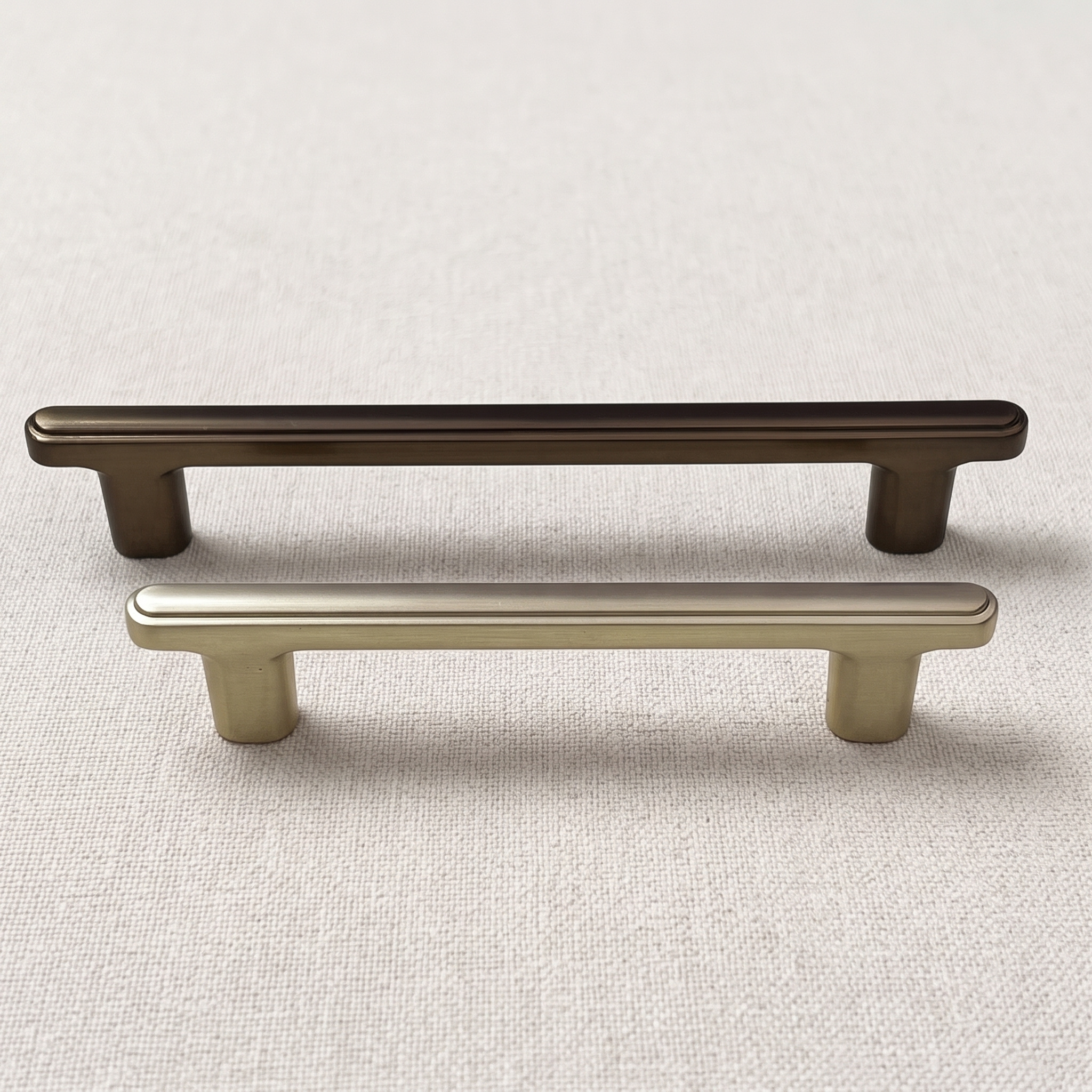 Two ridge-top bridge cabinet pulls in Brush Gold and Coffee resting on a pale linen surface