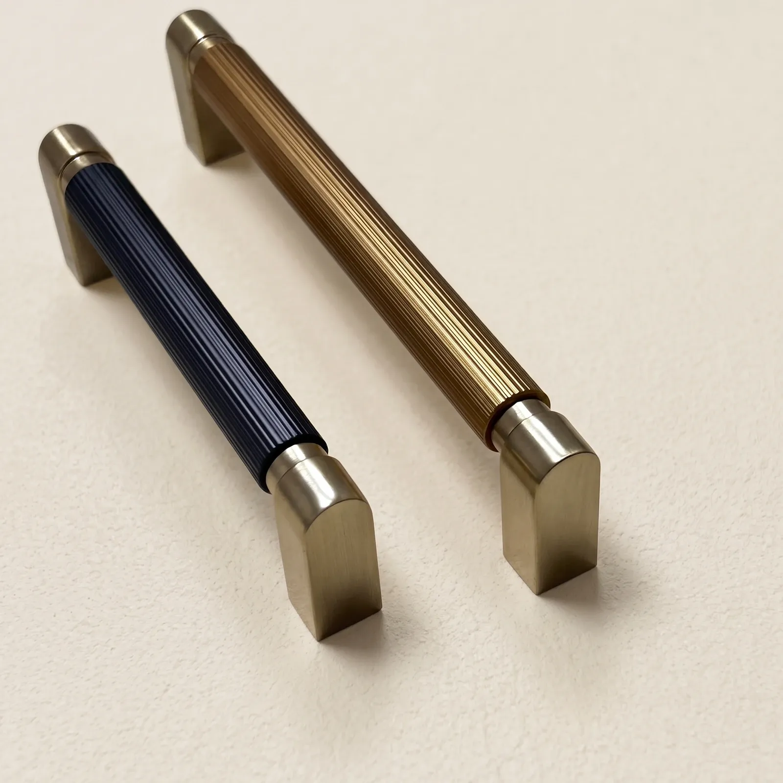 Two color-blocked D-profile pulls on a cream backdrop, one with a dark gray grooved grip and one in brass gold, both with brushed gold end blocks