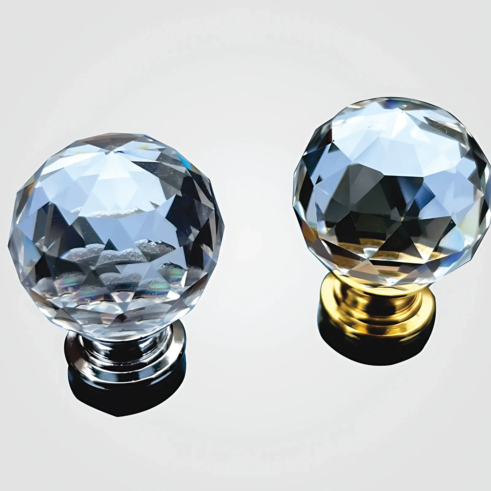 Angled view of two faceted clear crystal ball knobs side by side on bright ground; left on silver-tone base, right on gold-tone base.
