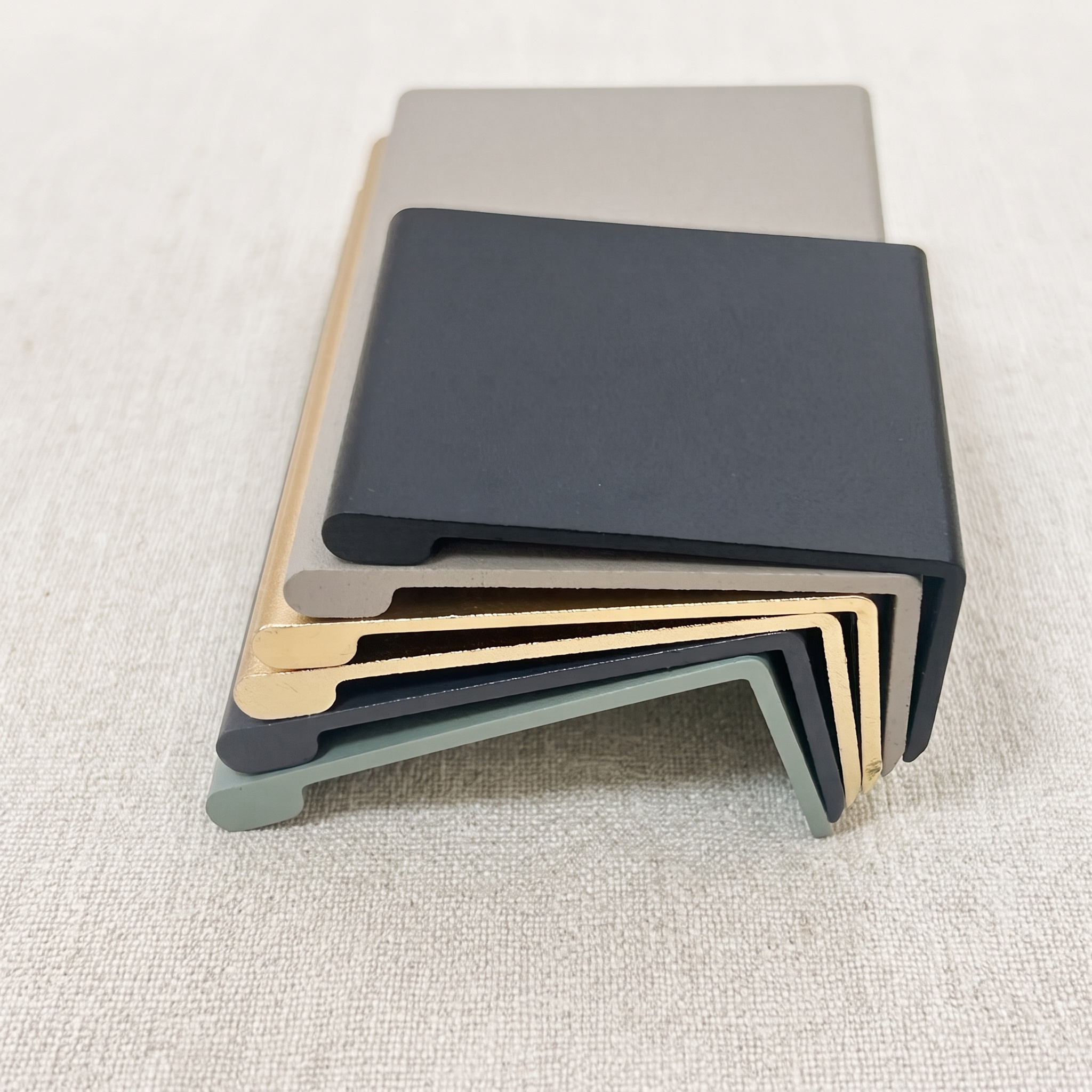 A stack of six smooth-arc edge pulls in khaki, champagne gold, matt black, gunmetal, and green finishes resting on a pale beige fabric surface.