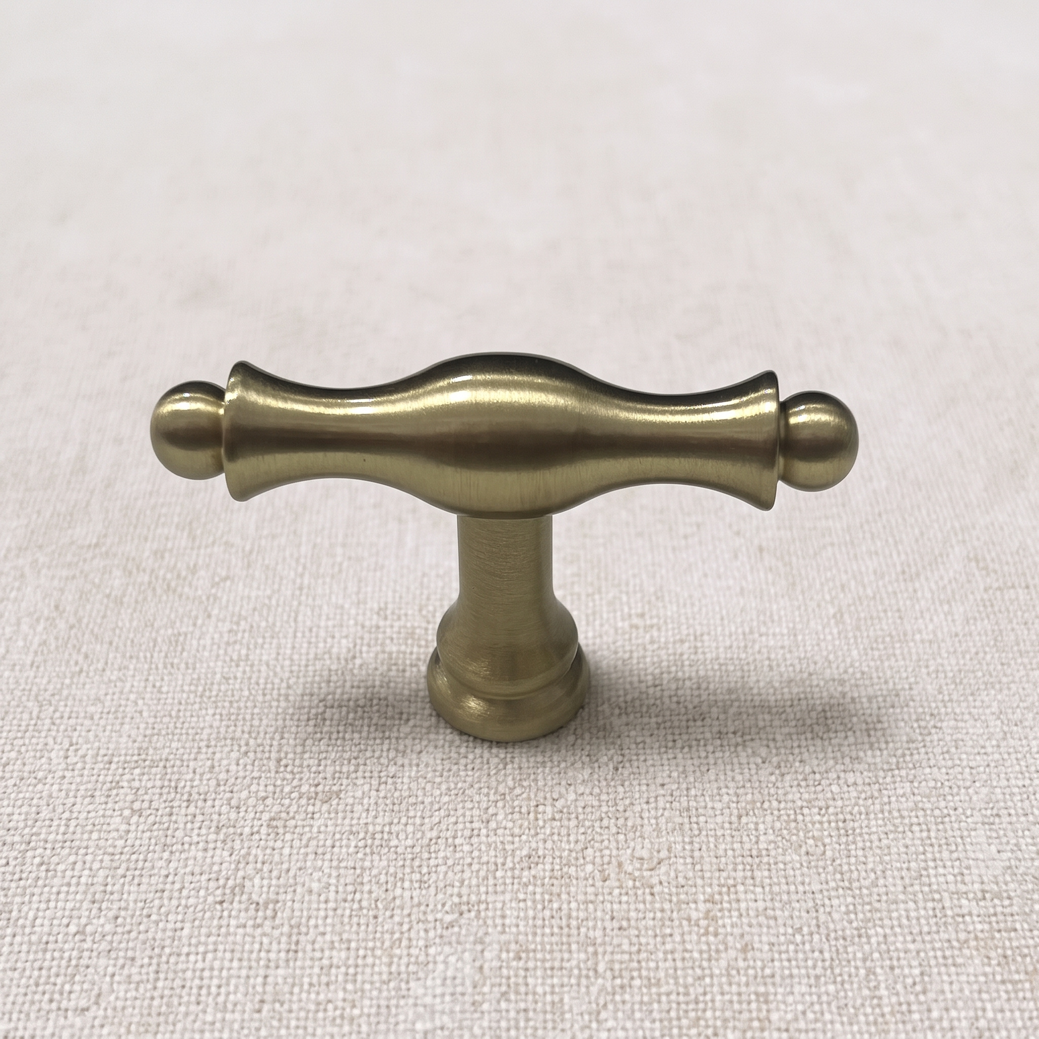 Front view of a Brass Gold wavy T-knob with a sinuous bar profile, ball-tipped ends and a stepped foot on a pale linen surface.