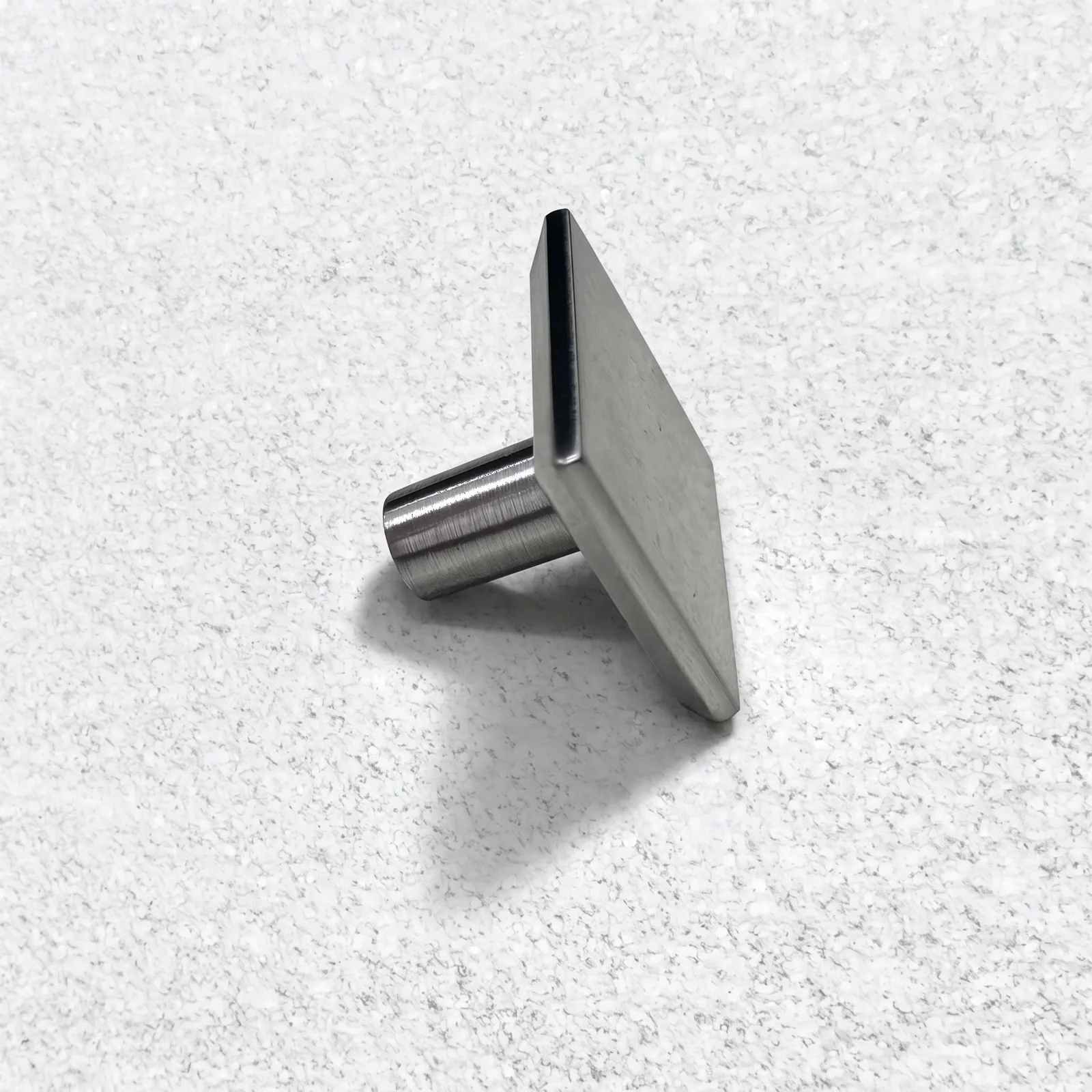 Brushed nickel square flat-plate cabinet knob seen from the side on a speckled ground.