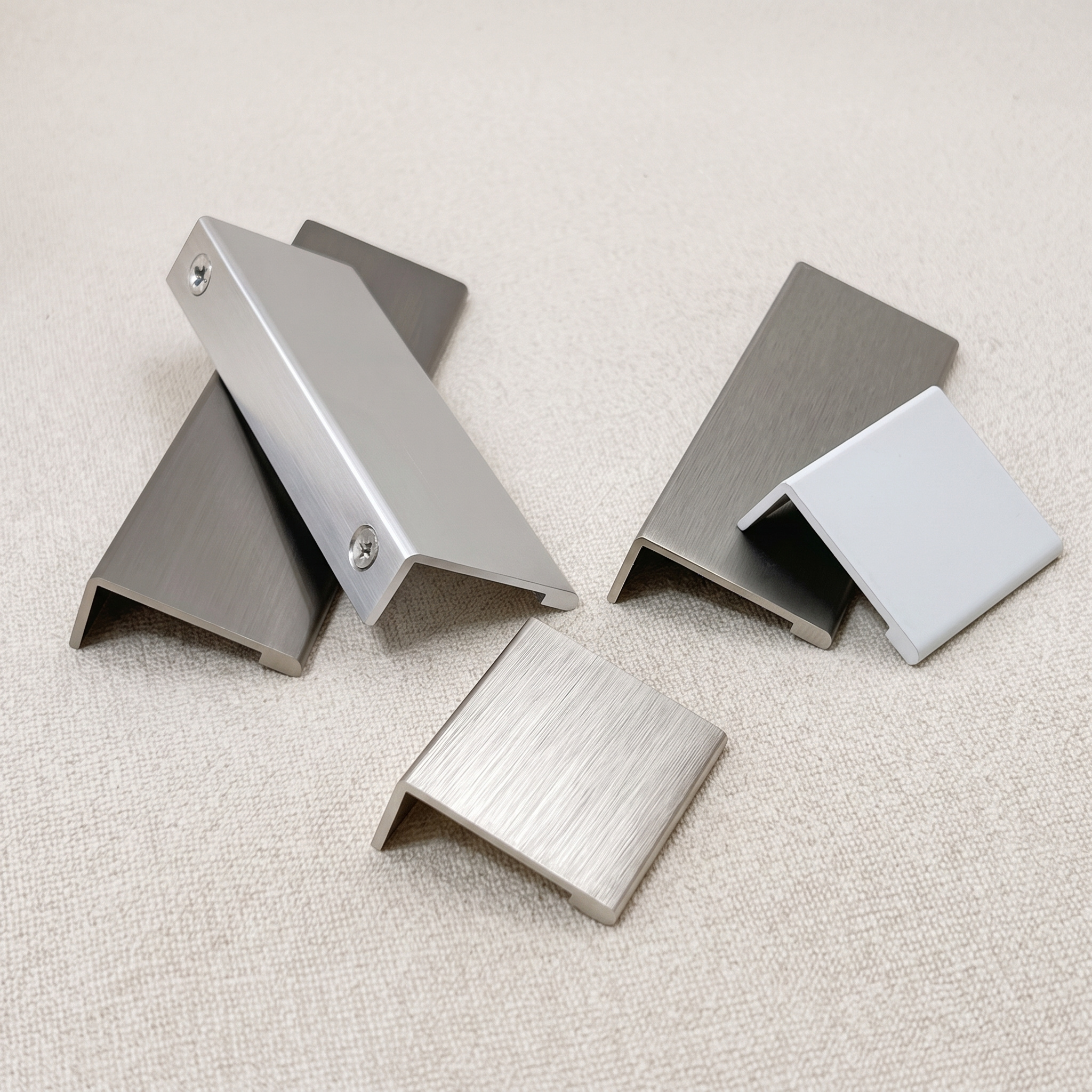 Close view of recessed L-profile edge pulls in matt black, chrome, brushed nickel, and white finishes on a linen surface.