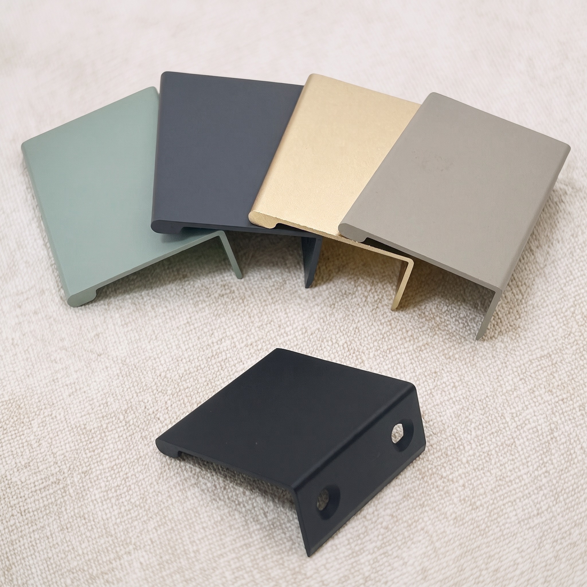 Five smooth-arc edge pulls on a pale beige fabric surface, with four fanned in green, gunmetal, champagne gold, and khaki behind a matt black pull that shows two countersunk holes.