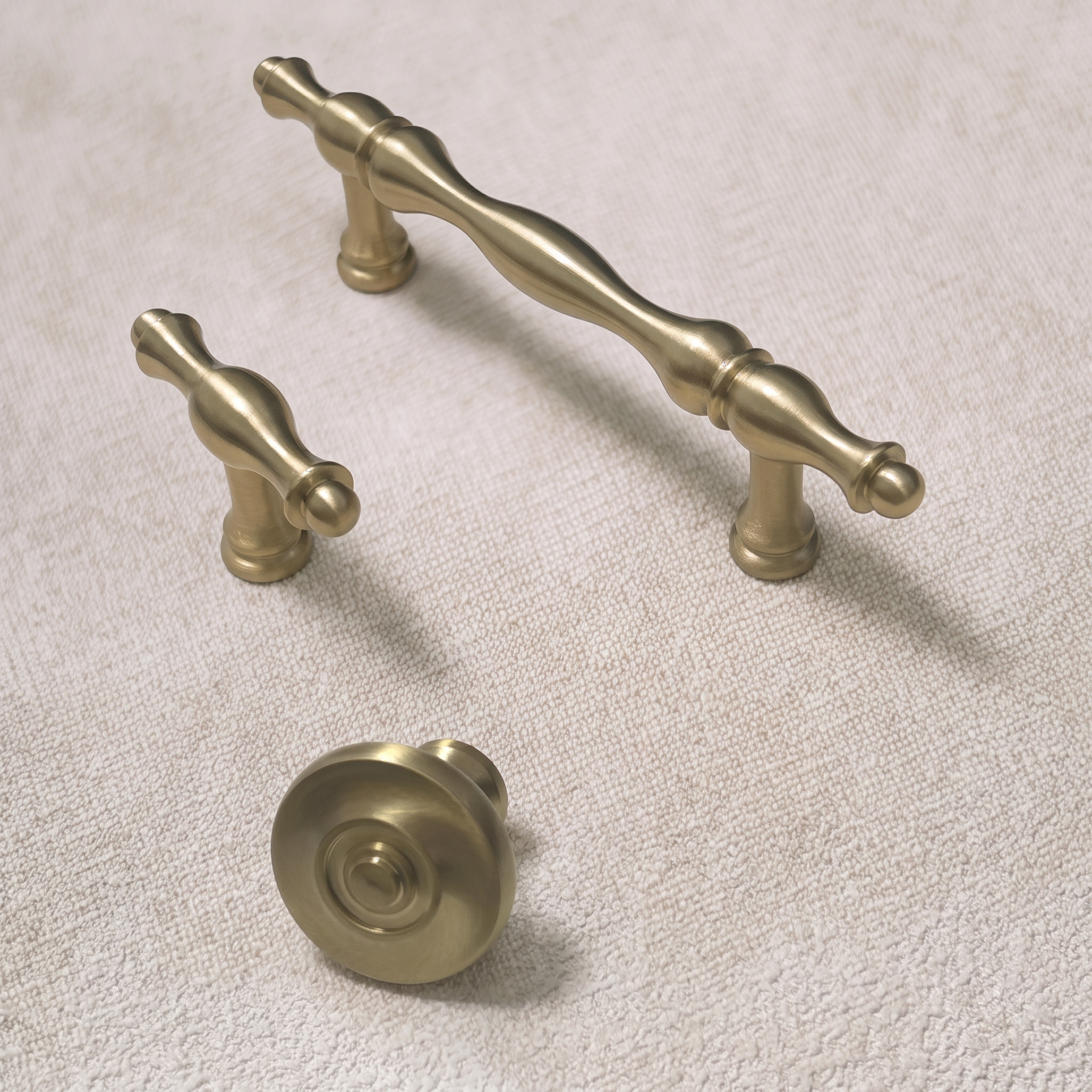 Angled view of the wavy brass family with the bar pull, T-knob and a round disc knob resting on its side to show the mounting post, on a pale linen surface.