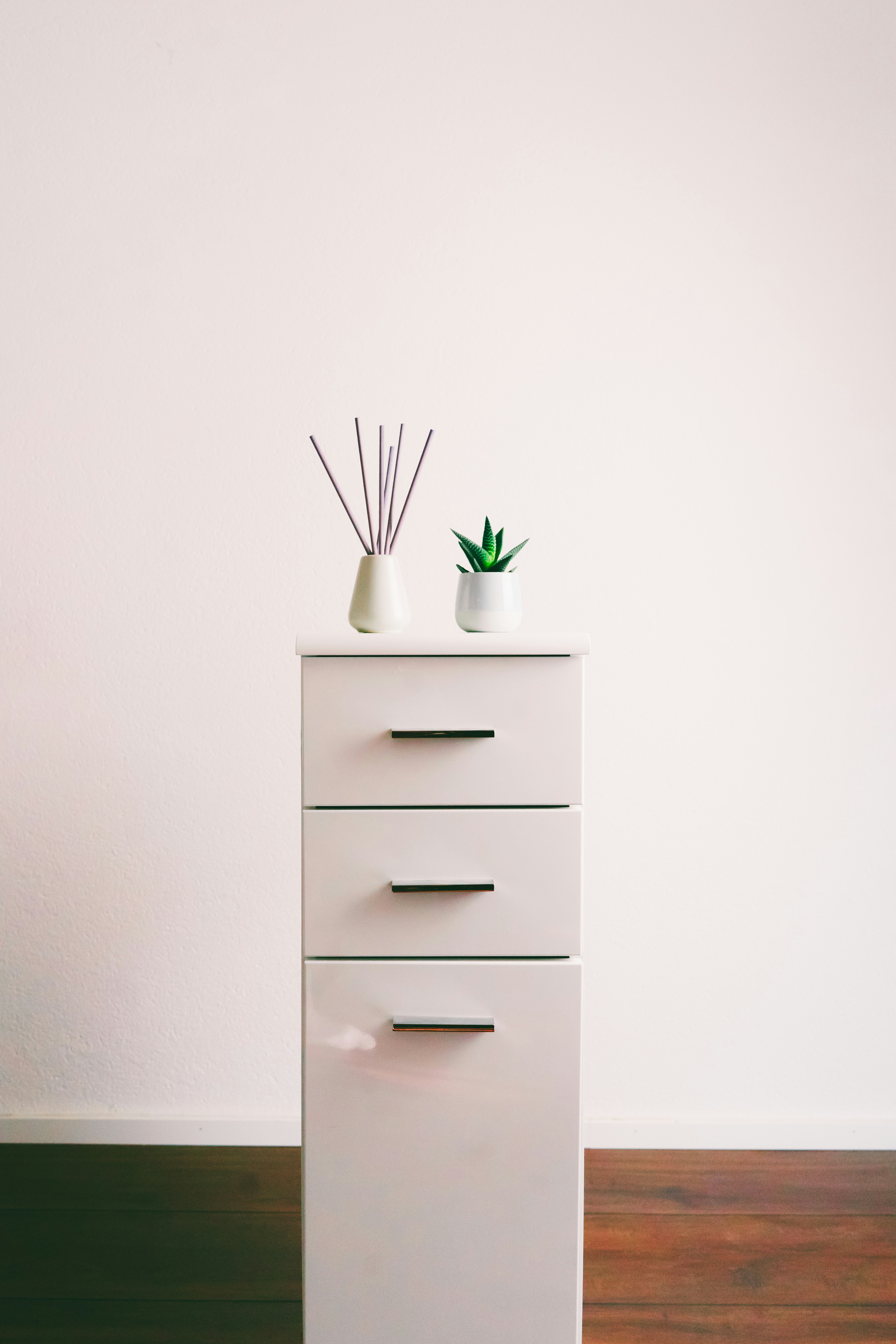 White chest of drawers with three slim dark drawer pulls, standing against a pale wall above a wood floor, with a diffuser bottle and a small potted plant on top.