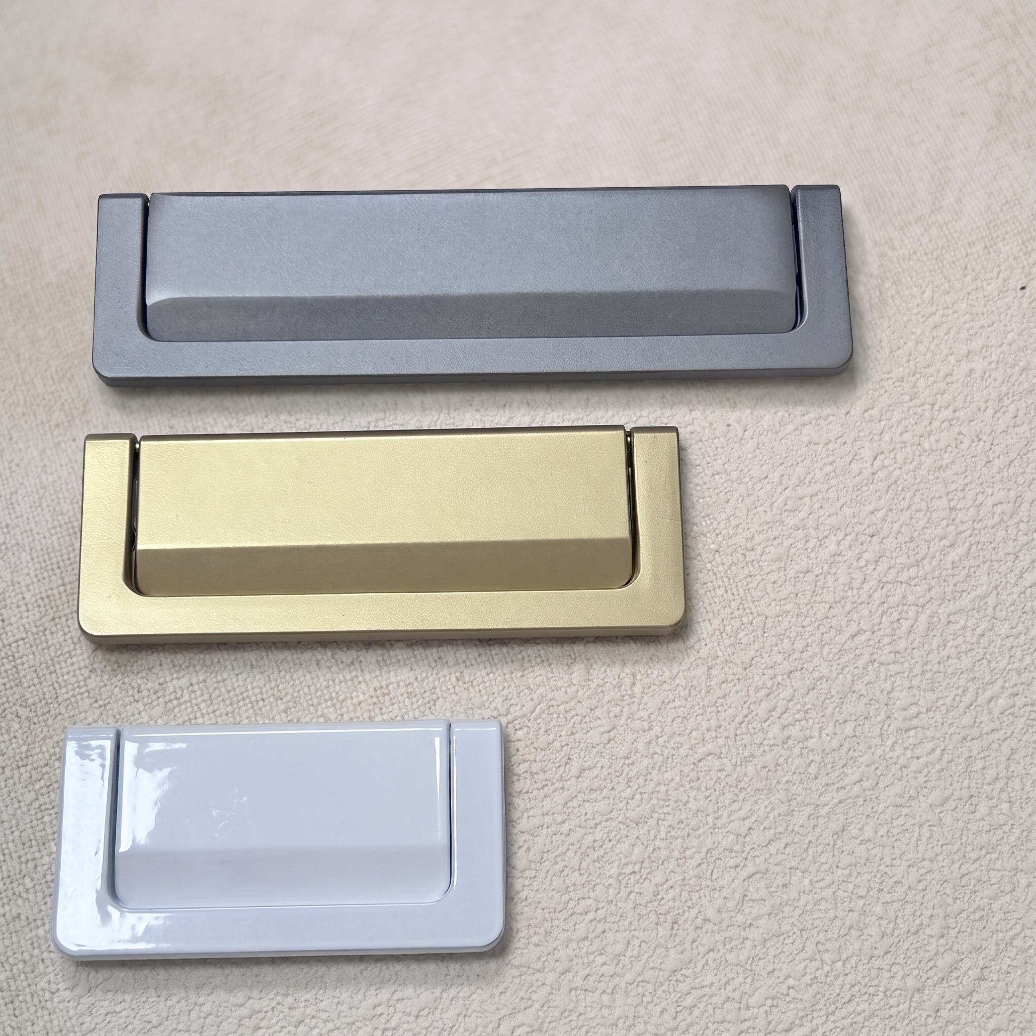 Three recessed flip pulls stacked in grey, brass gold and white, each with the tab flush with its plate, on a pale beige textured surface.