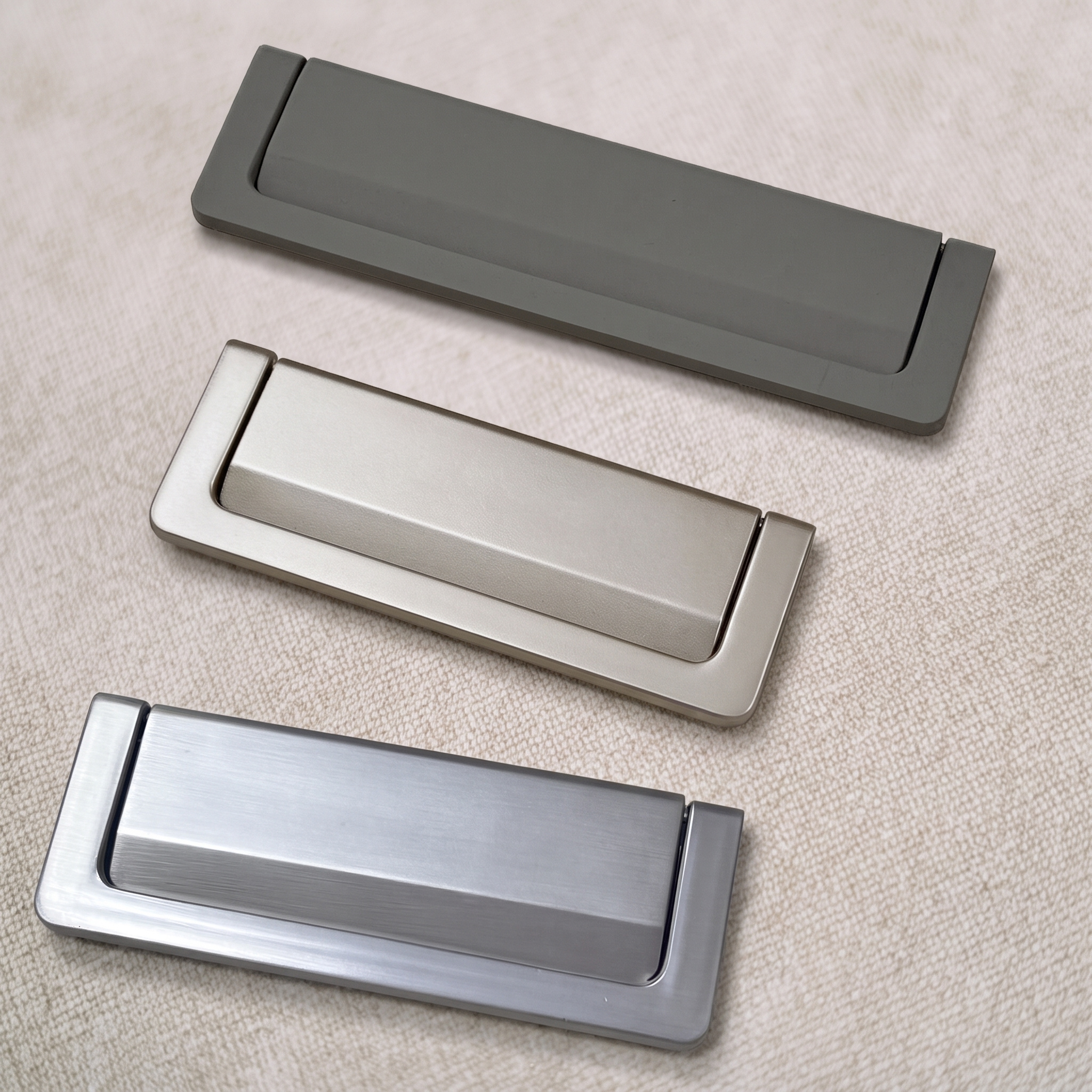 Three recessed flip pulls in graphite, champagne nickel and brushed chrome, arranged diagonally on a pale beige textured surface.