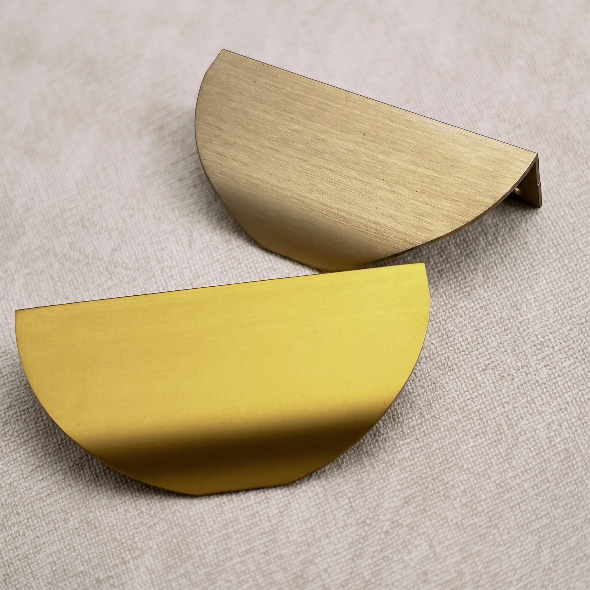 Two half-moon edge pulls resting on pale linen, one brushed warm gold and one bright gold, each showing its curved shell and rear lip