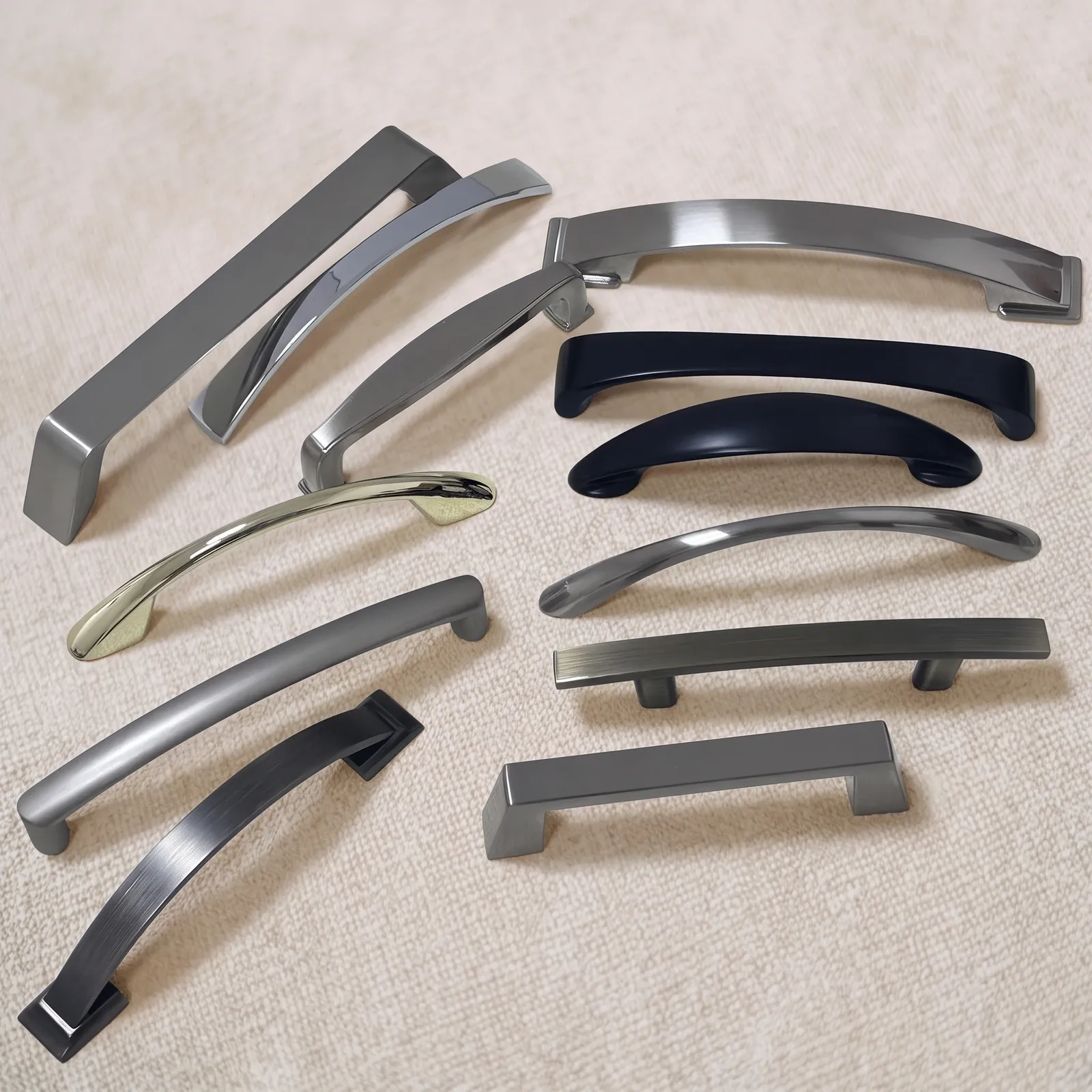 Group view of twelve cabinet handles from the series, mixing arched and straight bar profiles in matte black, gold-tone, silver-tone, dark bronze and pewter-grey finishes, arranged on a linen surface.