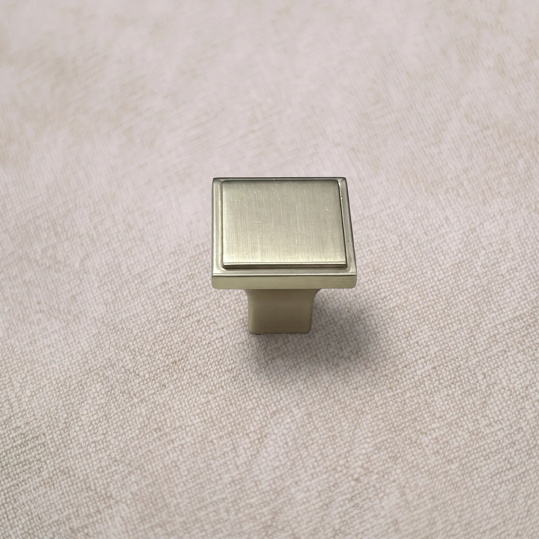 Angled view of a Brush Gold square cabinet knob showing its stepped rim, raised inner panel, and narrow pedestal on textured beige surface.