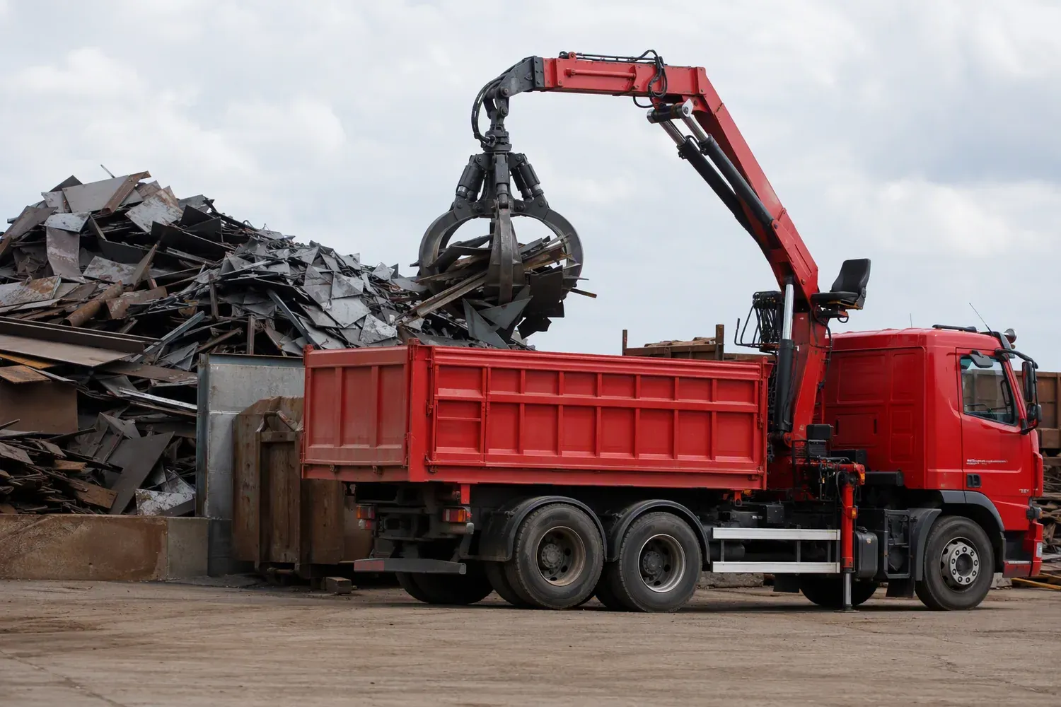 A truck-mounted grab truck lifting a bundle of cut steel plate with its knuckle-boom grab toward the open tipping body in a scrapyard