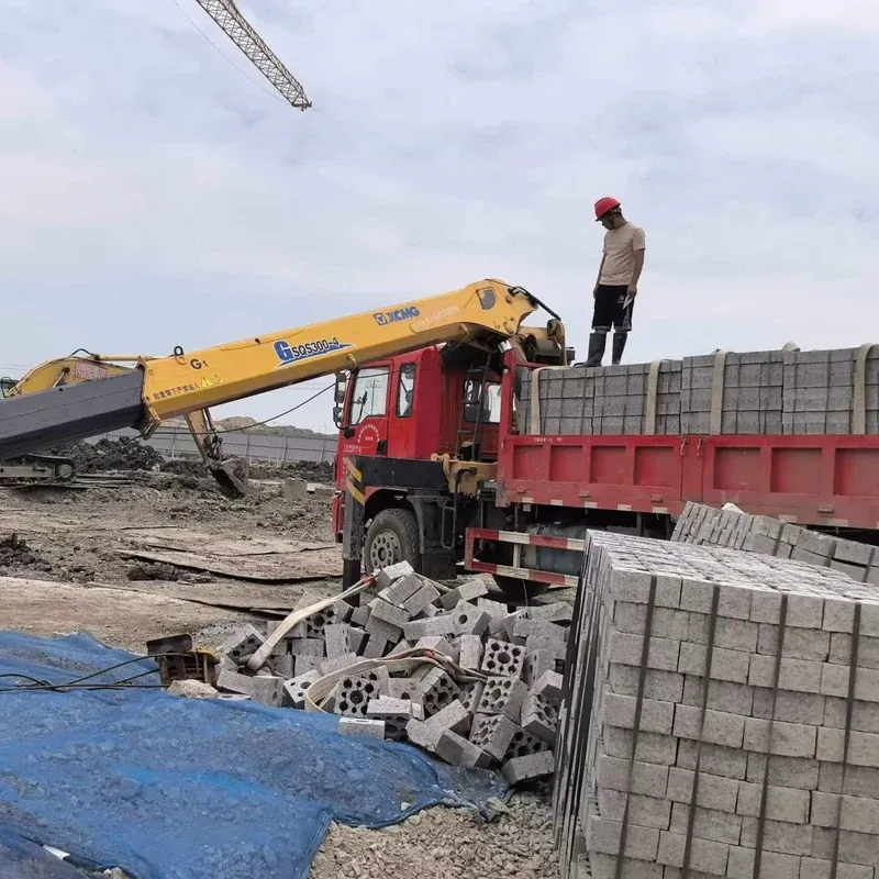 A truck-mounted crane working on a construction site with the boom extended past the truck, and a worker in a hard hat standing on the concrete block load