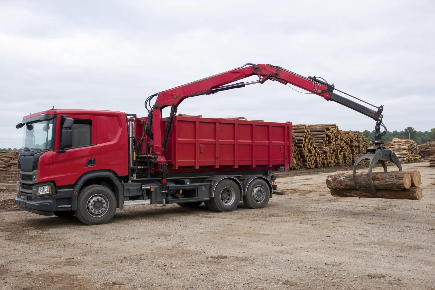 A truck-mounted grab truck with a knuckle-boom crane and log grapple holding two logs at a timber yard, dump body on the chassis