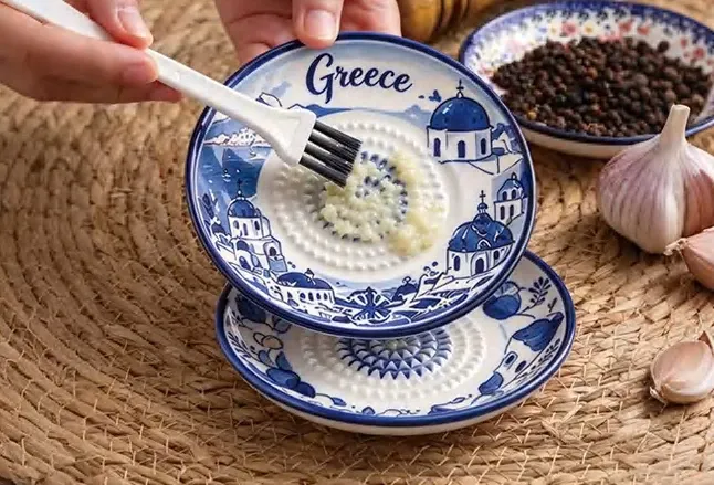 A hand using a spoon over a blue-and-white Greece design ceramic garlic grater dish with a textured center.