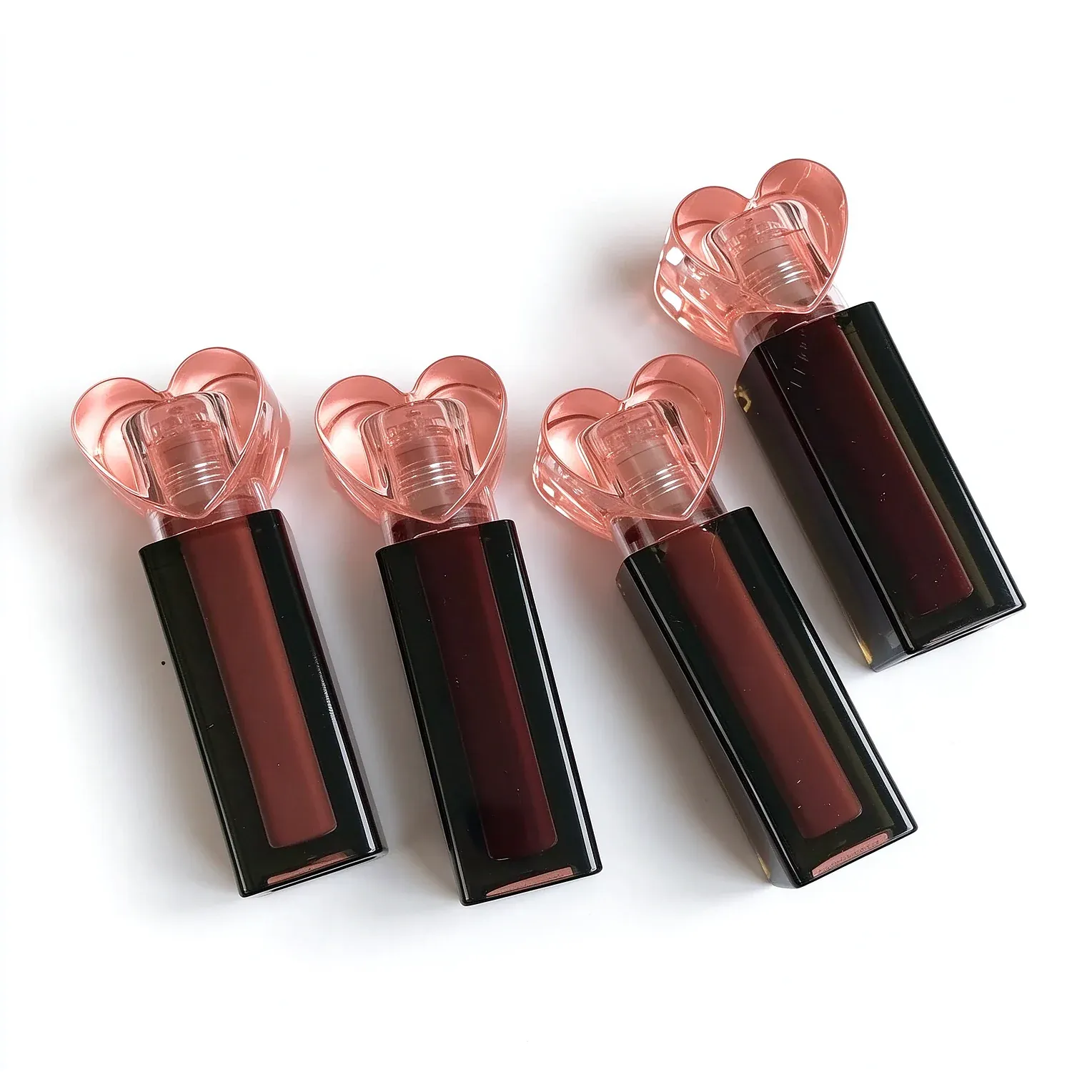 Four square black lip gloss bottles with clear heart-shaped caps in nude, rose and berry shades