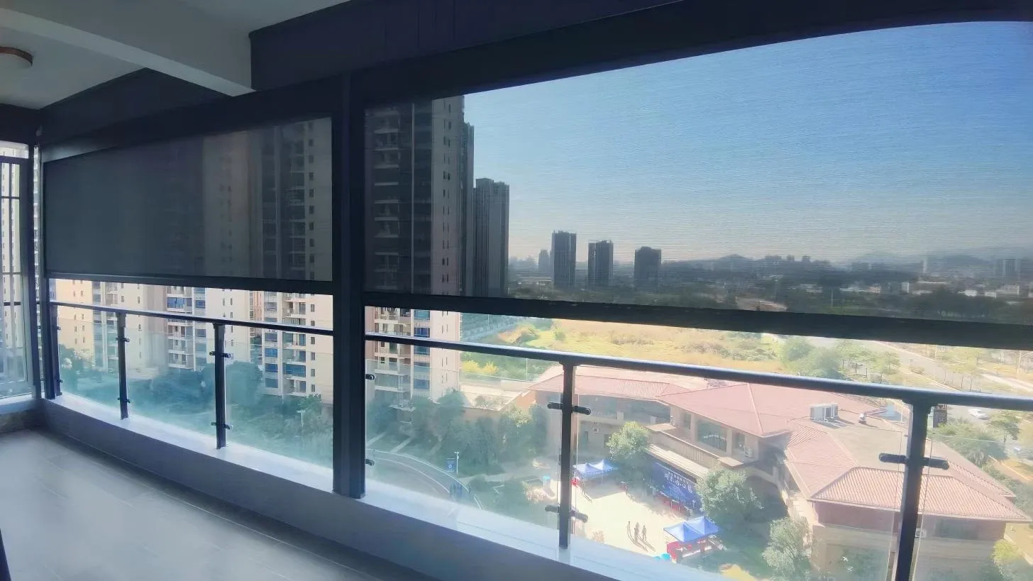 Motorized windproof roller blind fitted across a balcony opening, partially lowered, with a city view of buildings and hills beyond.