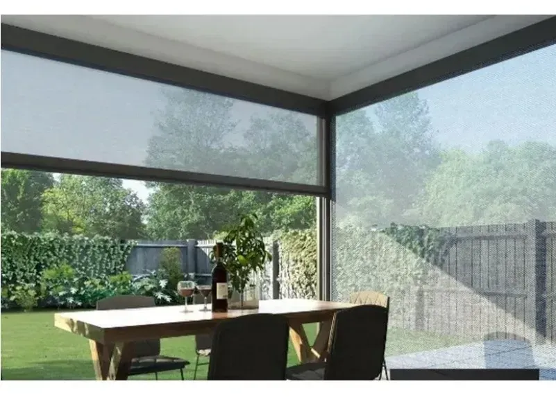 Hand Crank manual windproof roller blind fitted at an indoor dining area open to a garden, blind partially raised with sunlight shading the table.