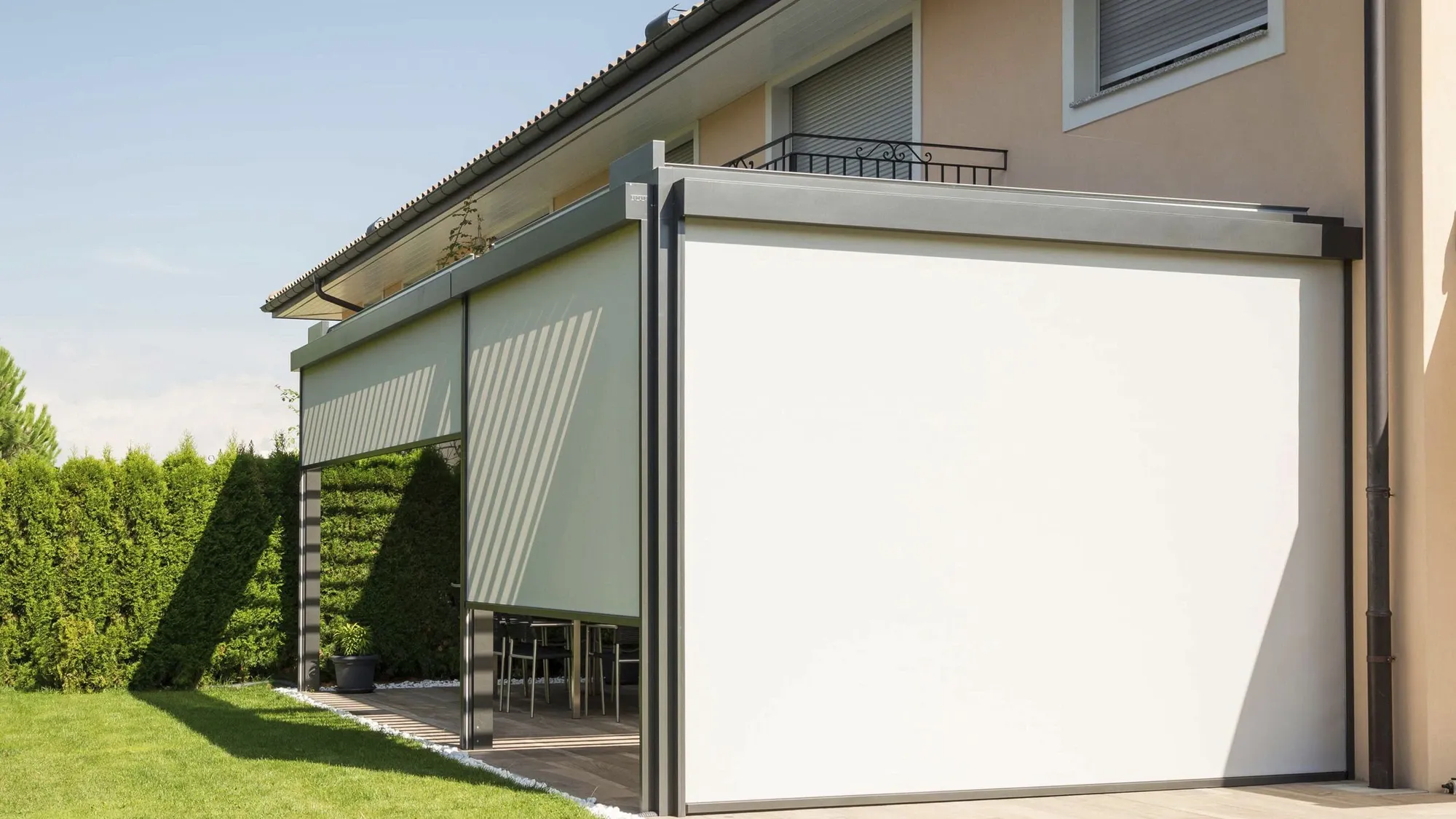 Manual windproof roller blinds on a covered house terrace, with one panel fully lowered and one half lowered in aluminium side tracks beside outdoor dining chairs, lawn and hedge.