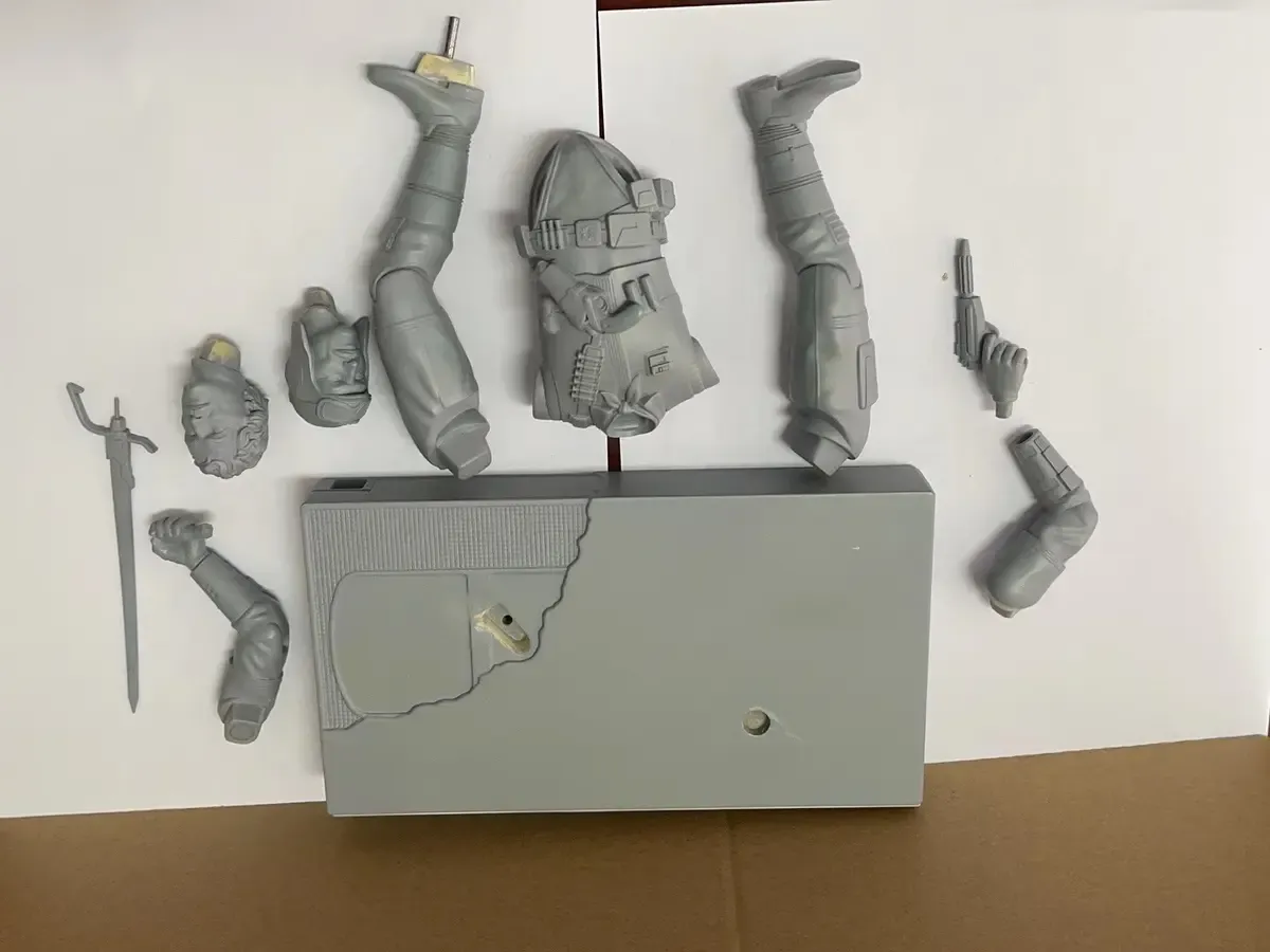 Unpainted gray collectible torso, limbs, head and accessories arranged beside a separate rectangular base with locating holes