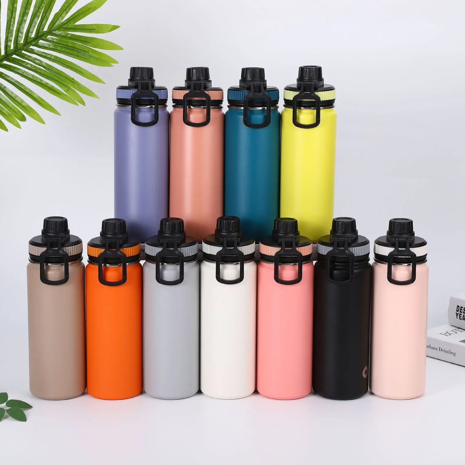 Group of eleven colorful spout-lid stainless steel bottles in assorted finishes arranged in a lifestyle scene with a palm leaf