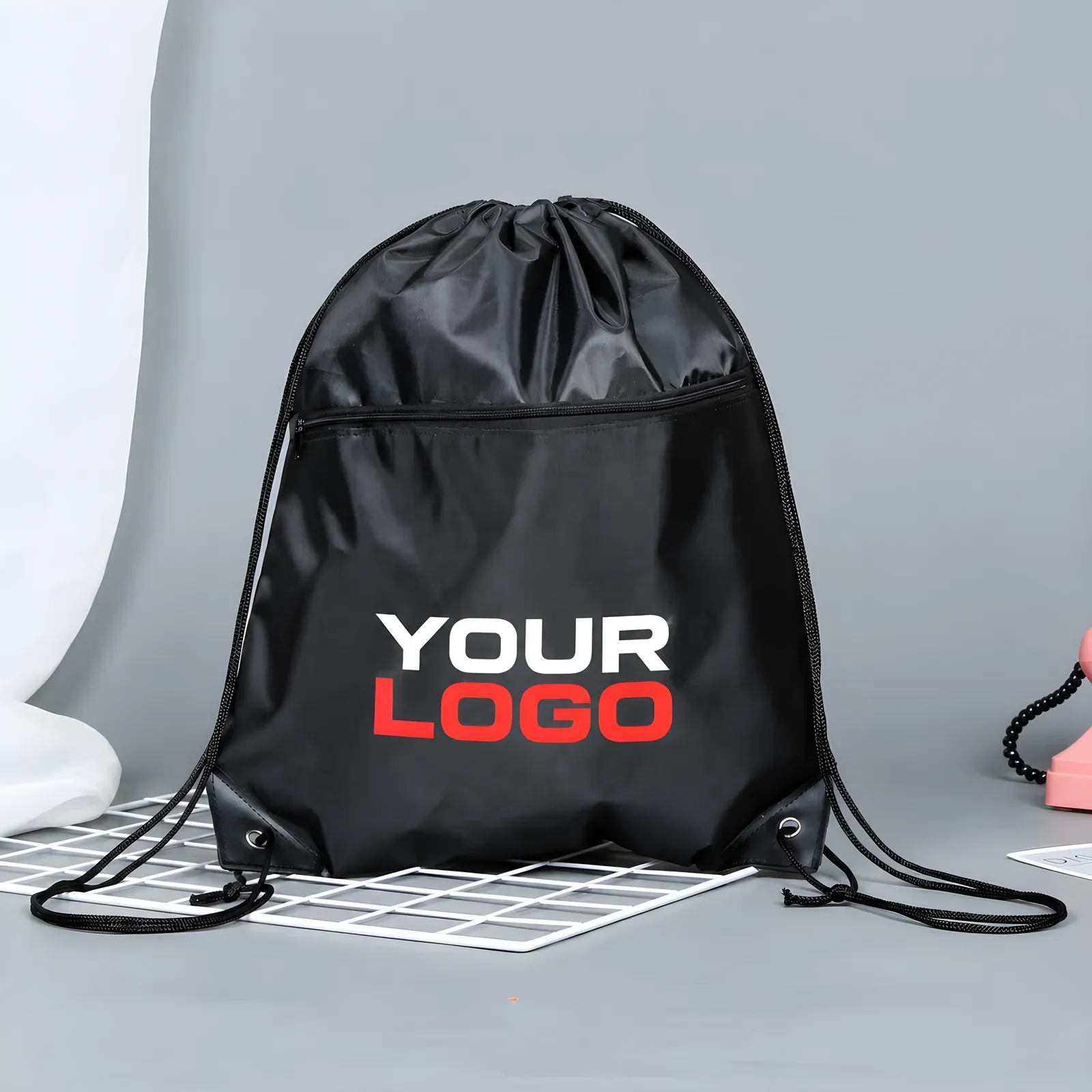 Black heavy-duty 3000D drawstring backpack with front zip pocket and YOUR LOGO print in white and red, on a grey studio background