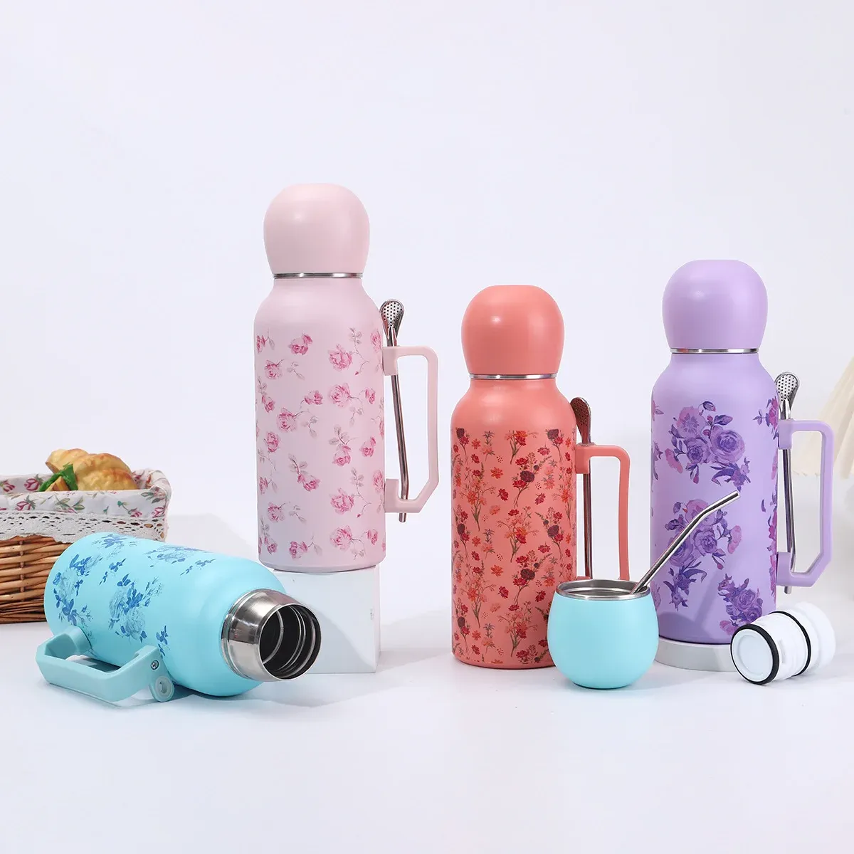 Four 1300ml floral vacuum bottles in pink, coral, purple and light blue with dome lids, side handles, metal straws and a small matching cup, lifestyle scene
