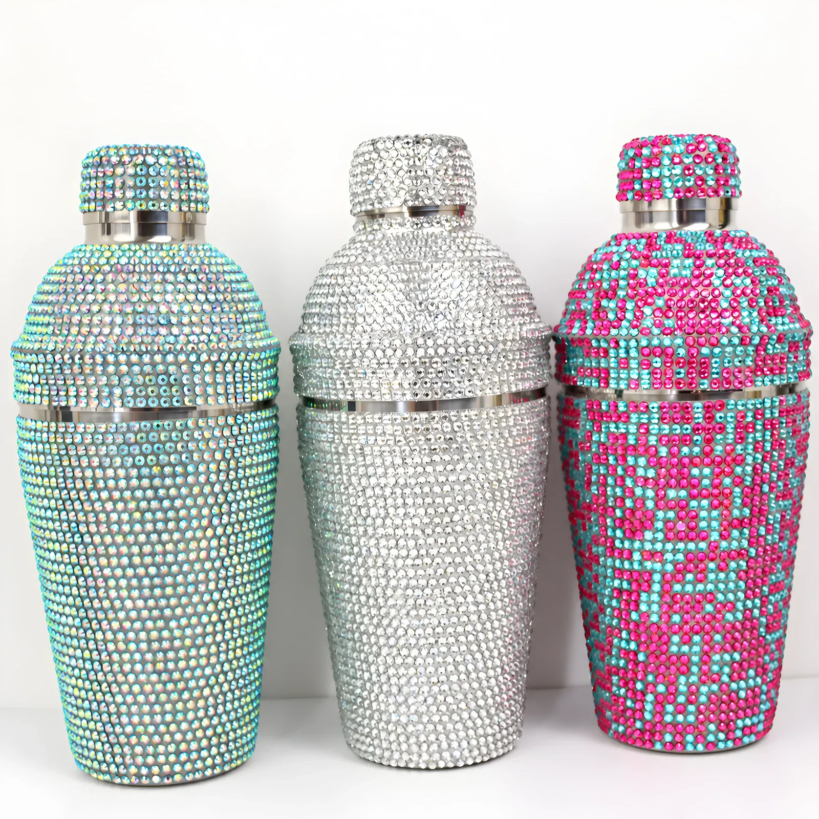 Three rhinestone-covered cocktail shakers in iridescent teal, clear and pink-teal finishes on a white background