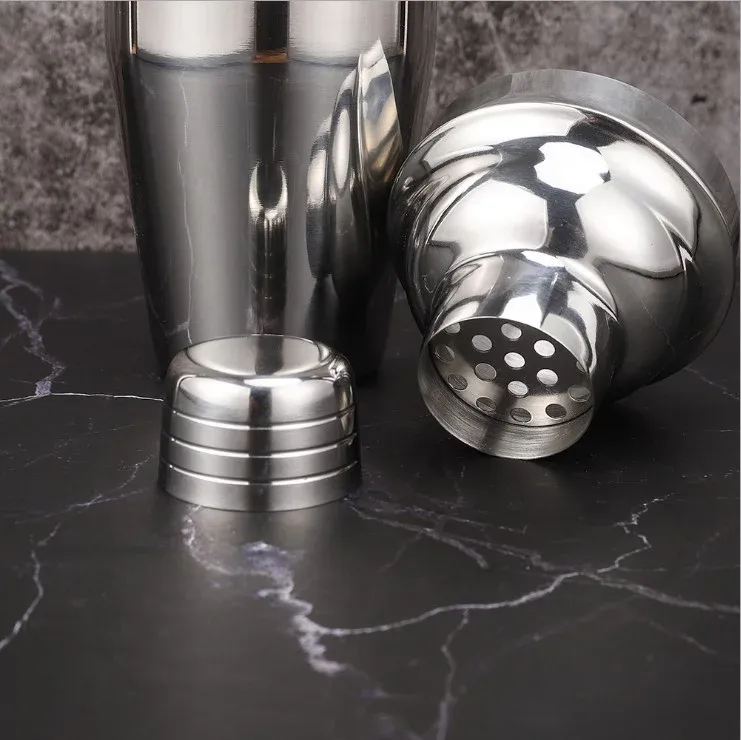Disassembled silver cocktail shaker showing body, strainer top and cap on a dark marble surface