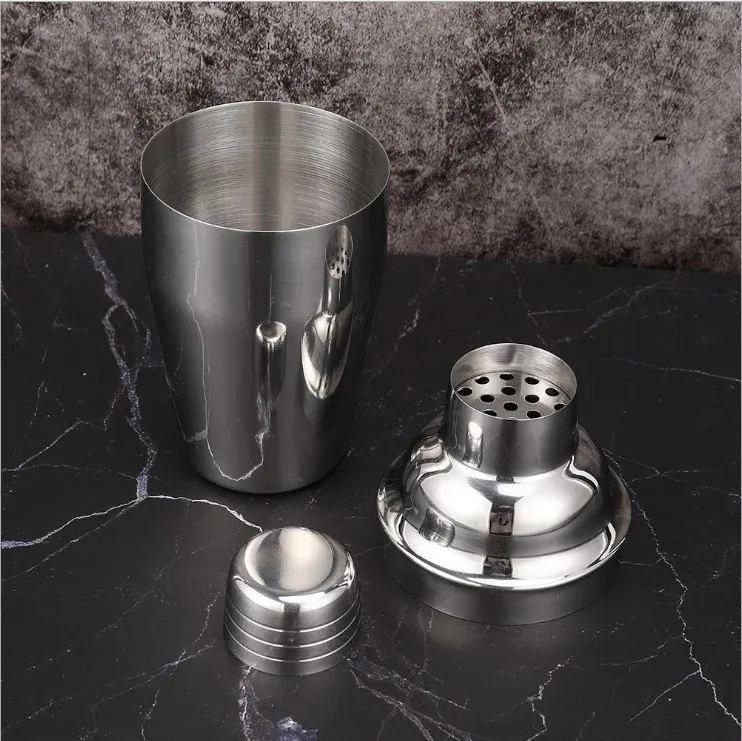 Silver cocktail shaker taken apart into body, strainer top and small cap on a dark marble surface
