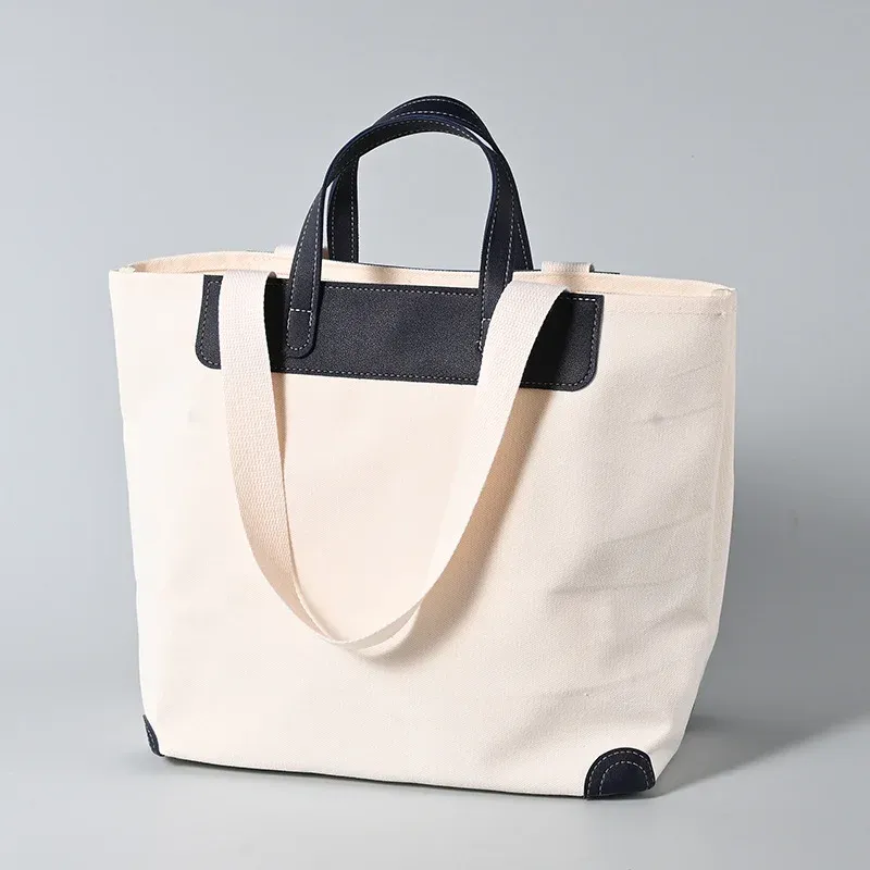 Ivory 16oz canvas tote bag with navy leather short handles, trim panel and corner guards, long webbing shoulder handle draped in front