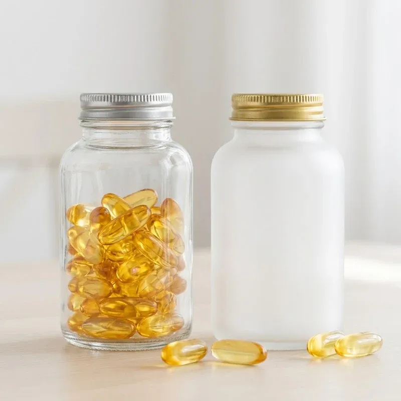 Clear glass capsule bottle with silver cap filled with golden softgel capsules beside a frosted bottle with gold cap