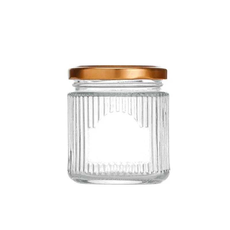 Squat clear glass jar with fluted ribbed body, arched label window and flat gold metal screw lid on white background